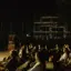 Pubblico seduto all'aperto di notte, che assiste a uno spettacolo o a un evento. Sullo sfondo si vede un edificio in costruzione, parzialmente completato e dotato di impalcature.