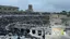 Un grande anfiteatro antico all'aperto gremito di spettatori. Sul palco si esibiscono artisti in costumi colorati. Sullo sfondo sono visibili delle rovine.