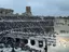 Un grande anfiteatro antico all'aperto gremito di spettatori. Sul palco si esibiscono artisti in costumi colorati. Sullo sfondo sono visibili delle rovine.