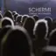 Una folla di persone rivolta verso un cartellone blu con la scritta "SCHERMI CINEMA MULTIPIAZZA", con un'illuminazione soffusa sullo sfondo.