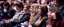 Audience seated in a theater, focused on an event. People of varying ages are engaged, seated in red chairs, with a blurred background.