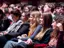 Spettatori seduti in un teatro, che guardano attentamente uno spettacolo o una presentazione, con espressioni concentrate e coinvolte.