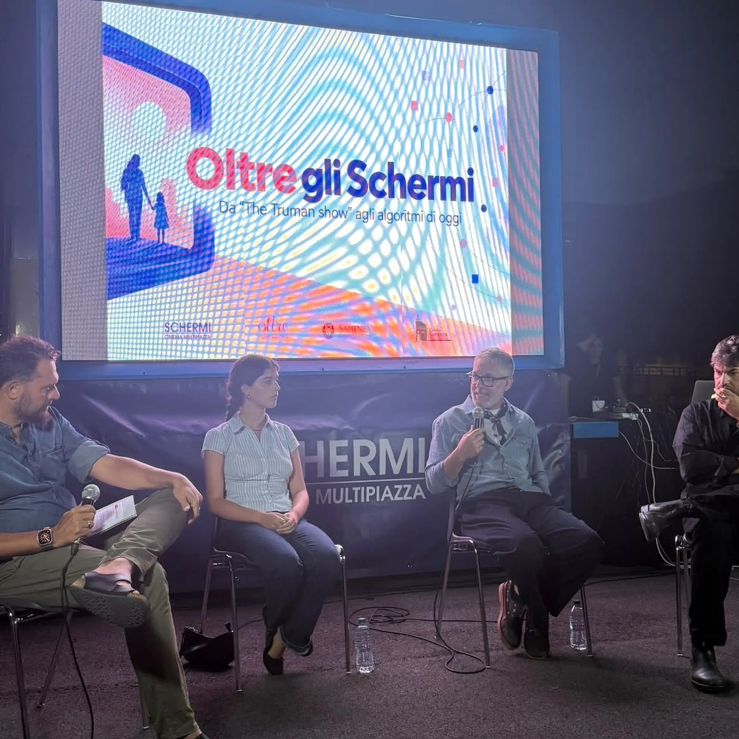 Quattro persone sedute su un palco durante una discussione, con un grande schermo alle spalle che mostra la scritta "Oltre gli Schermi" e altro testo in italiano.