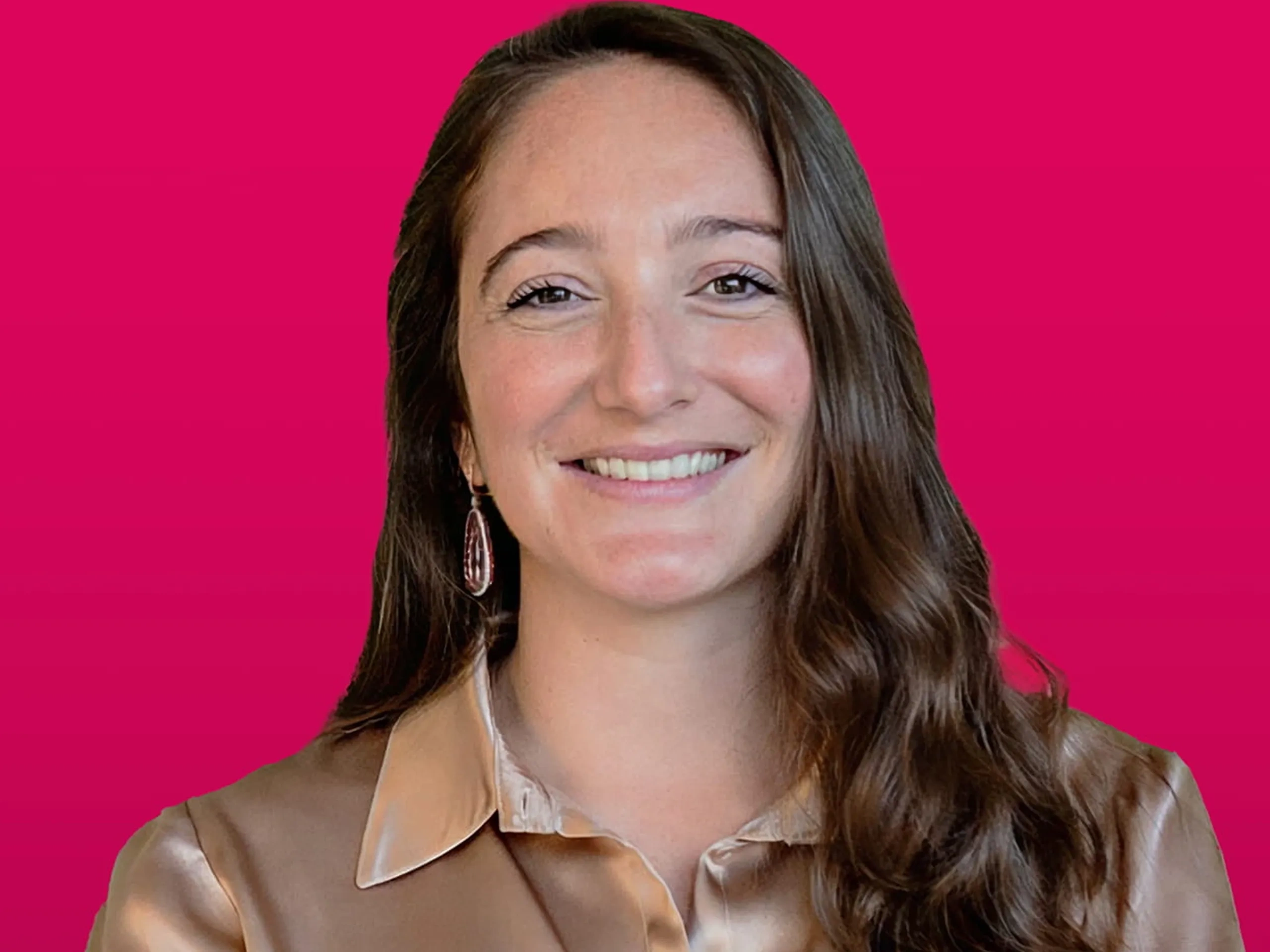 Donna sorridente con lunghi capelli castani, indossa una blusa color bronzo setosa, ha le braccia incrociate e si trova su uno sfondo rosa vivace.