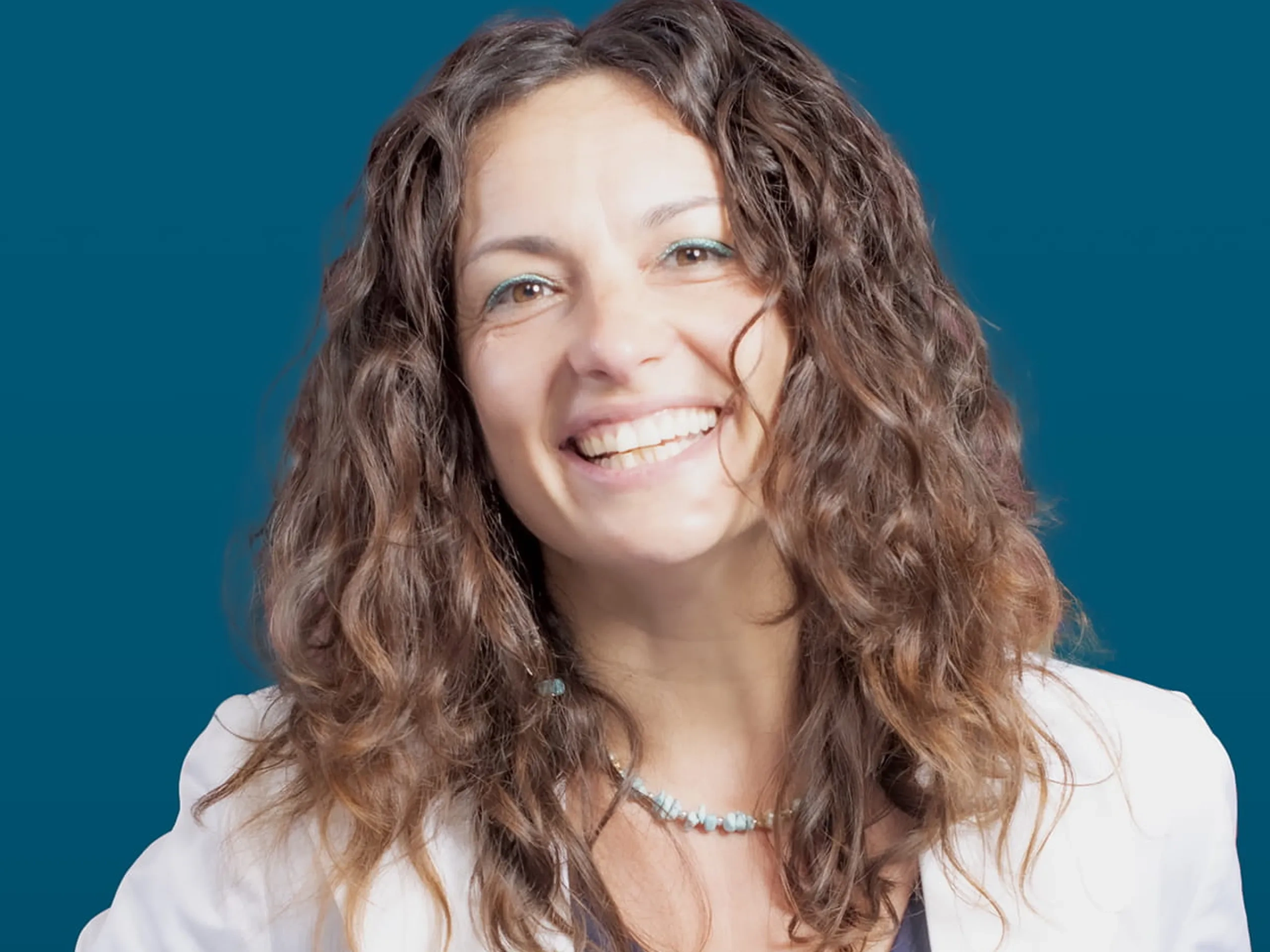 Donna sorridente con capelli castani mossi, che indossa un blazer bianco e una camicia blu scuro, in piedi su uno sfondo blu.