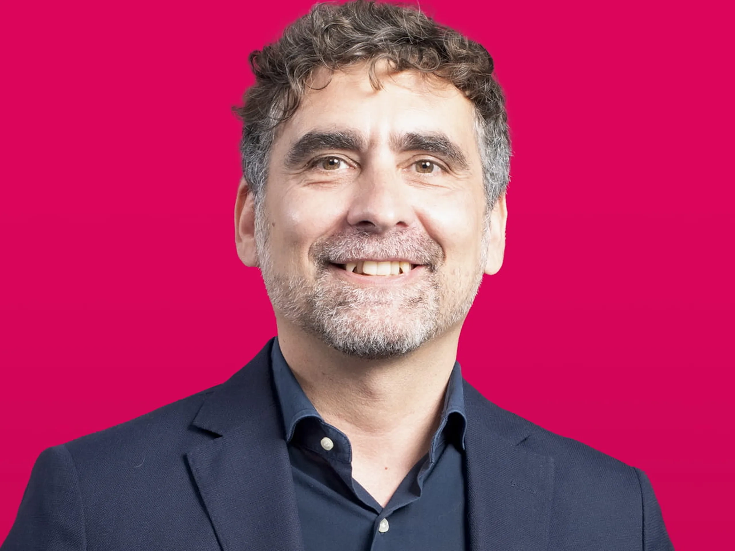 Uomo di mezza età sorridente con capelli ricci grigi e barba, con giacca blu scuro e camicia scura su uno sfondo magenta brillante.