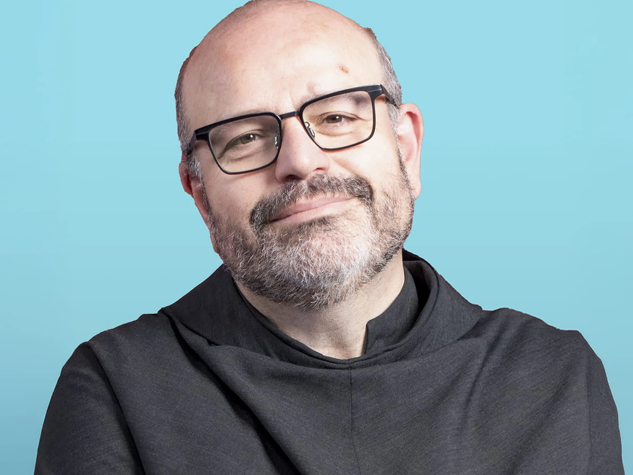 Uomo sorridente con occhiali e barba, che indossa un cappotto nero su uno sfondo azzurro.