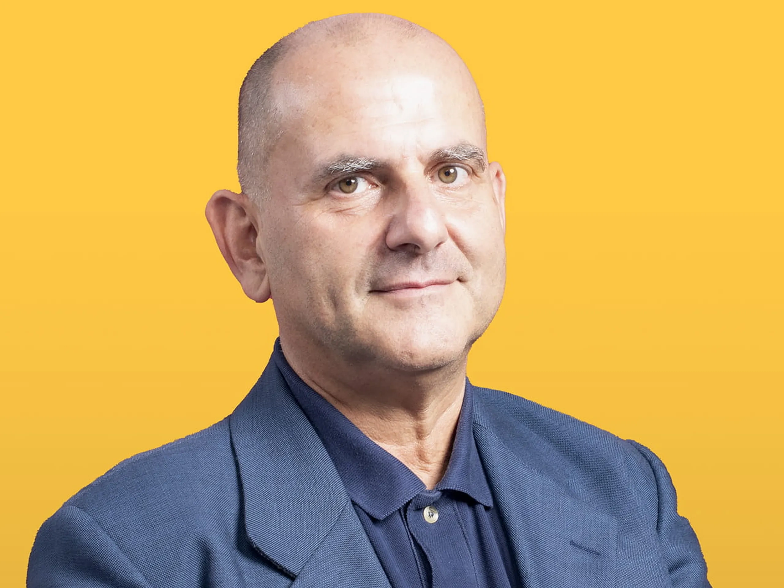Uomo calvo con giacca blu scuro e polo, mani giunte, espressione neutra su sfondo giallo.