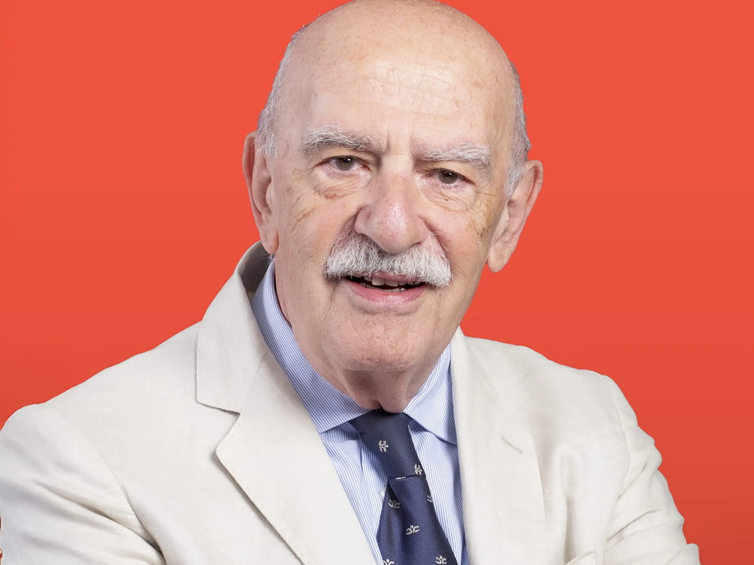 Anziano con baffi bianchi, vestito con un completo chiaro e cravatta a pois, in piedi con le braccia incrociate su uno sfondo arancione brillante.