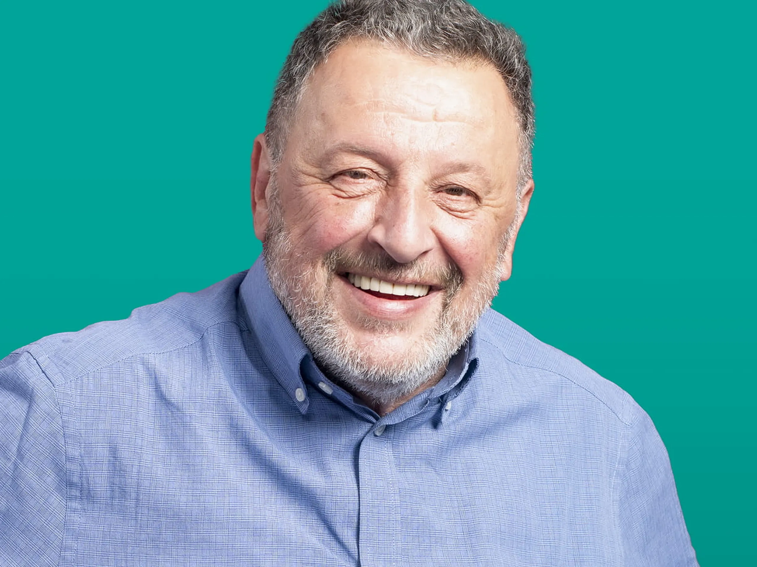 Uomo maturo sorridente con la barba, che indossa una camicia azzurra abbottonata, con una mano sul fianco, su uno sfondo verde acqua.