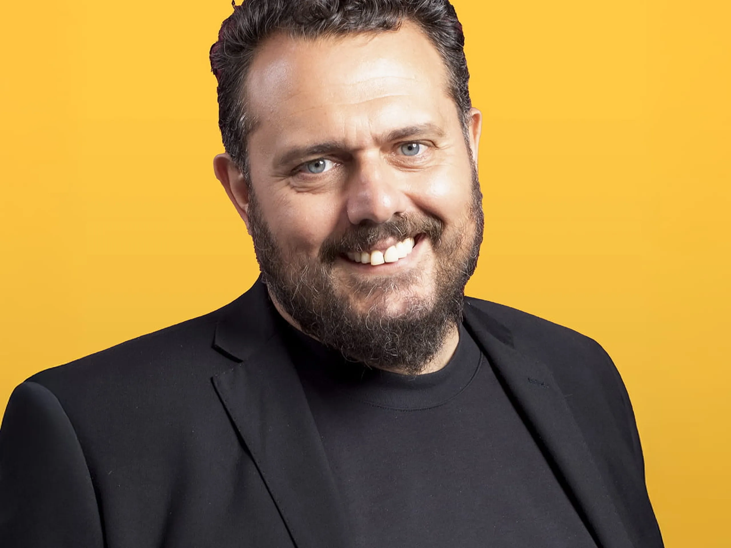 Uomo sorridente con la barba, che indossa un blazer nero e una camicia, in piedi su uno sfondo giallo brillante.