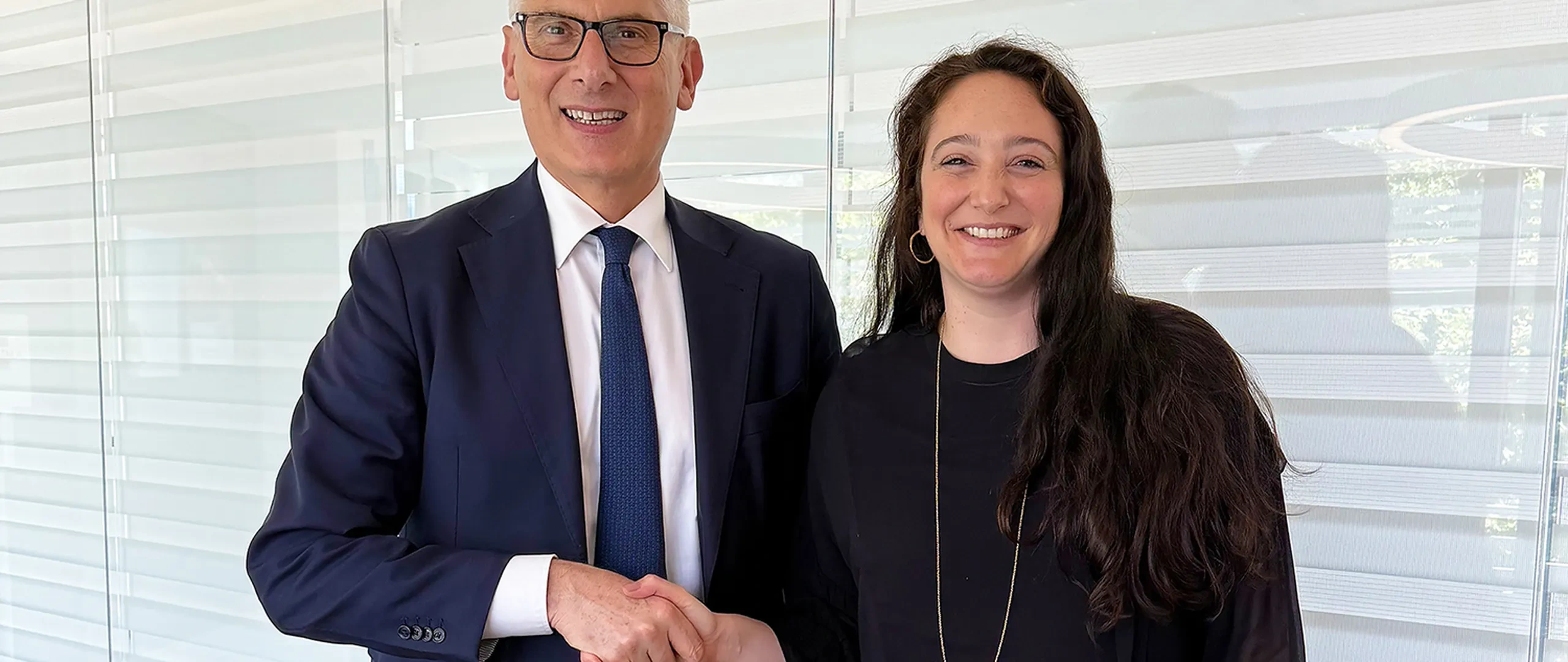 Uomo sorridente in giacca e cravatta e donna in abito nero che si stringono la mano davanti alle tende della finestra.