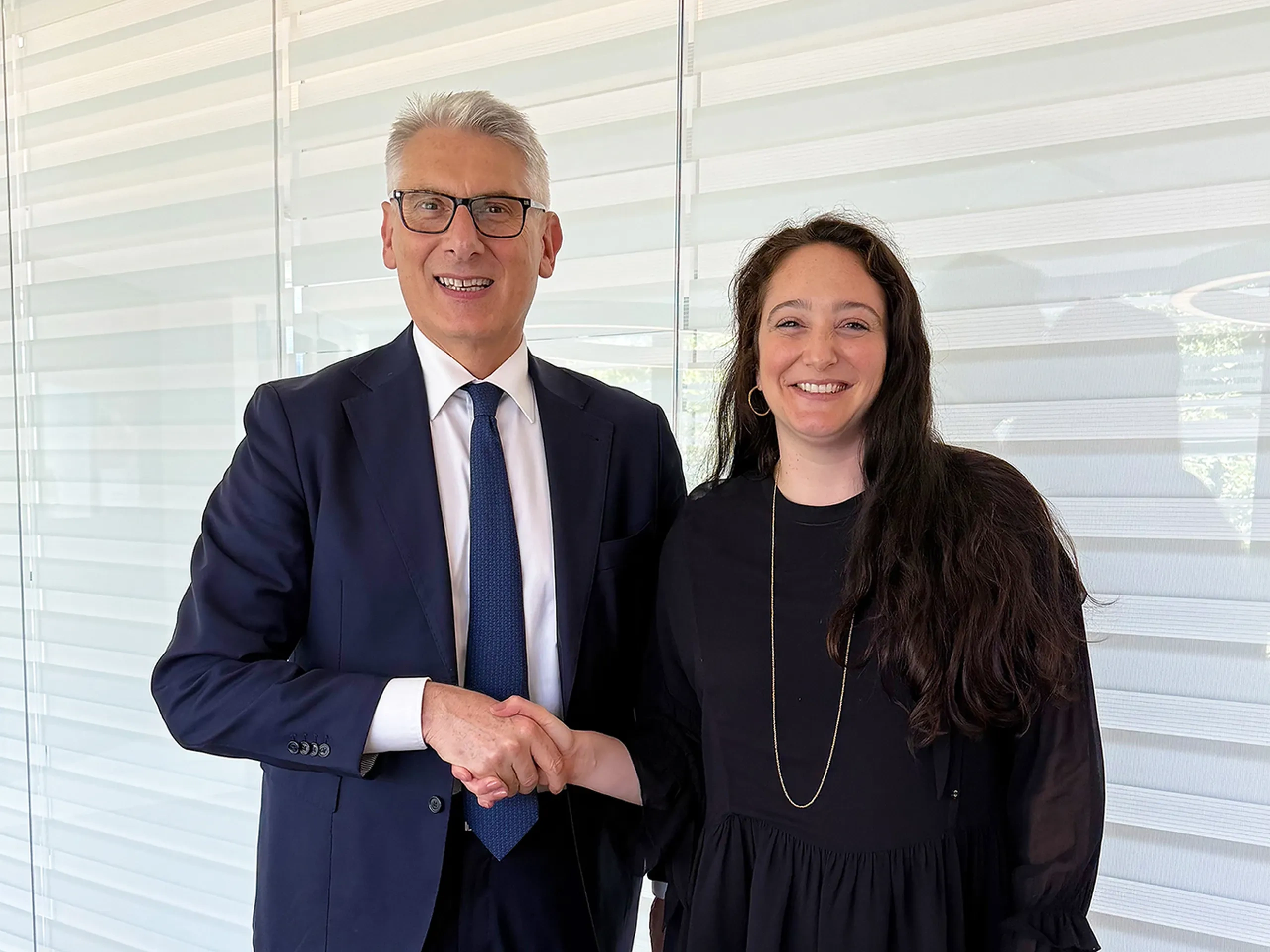 A man in a suit and a woman in a black dress smiling and shaking hands in front of a window with blinds.