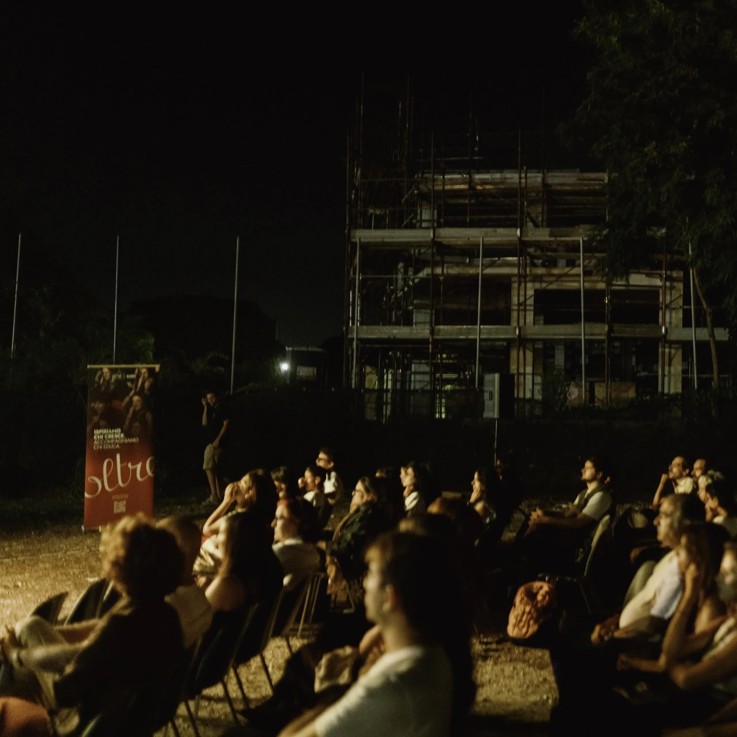 Pubblico seduto all'aperto di notte, che assiste a uno spettacolo o a un evento. Sullo sfondo si vede un edificio in costruzione, parzialmente completato e dotato di impalcature.