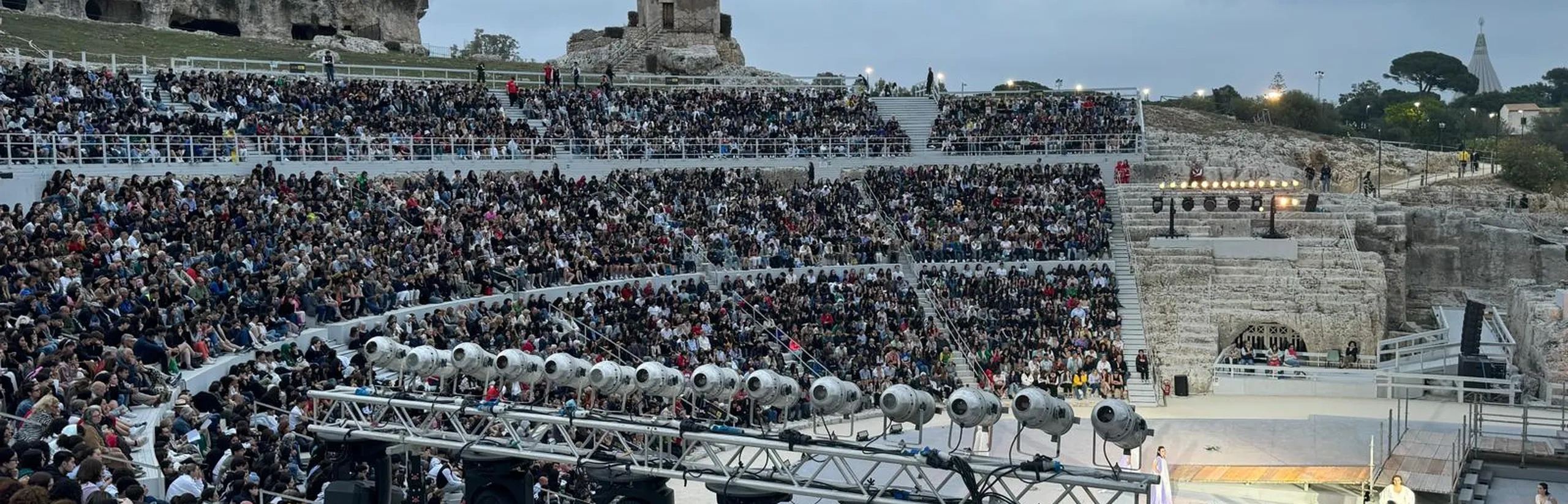 Un grande anfiteatro antico all'aperto gremito di spettatori. Sul palco si esibiscono artisti in costumi colorati. Sullo sfondo sono visibili delle rovine.