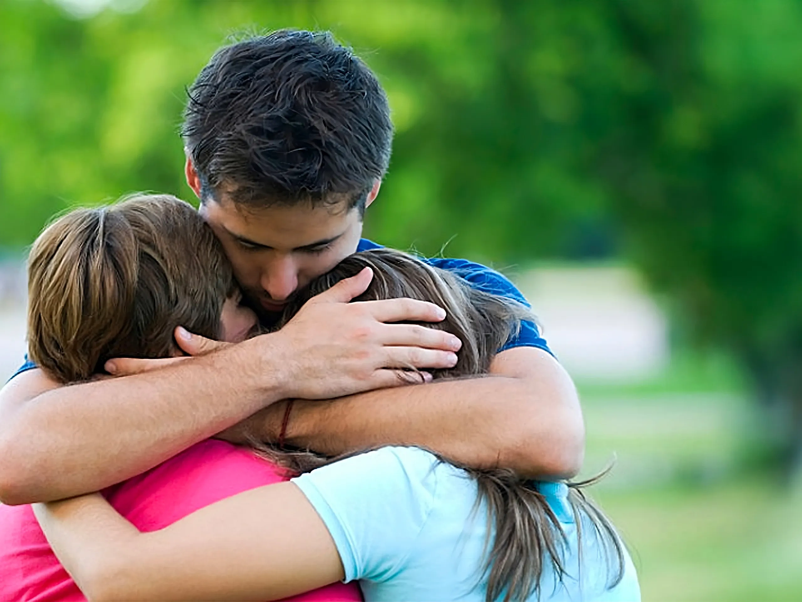 Tre persone si abbracciano all'aperto, circondate dal fogliame verde. Sembrano unite e rassicuranti.