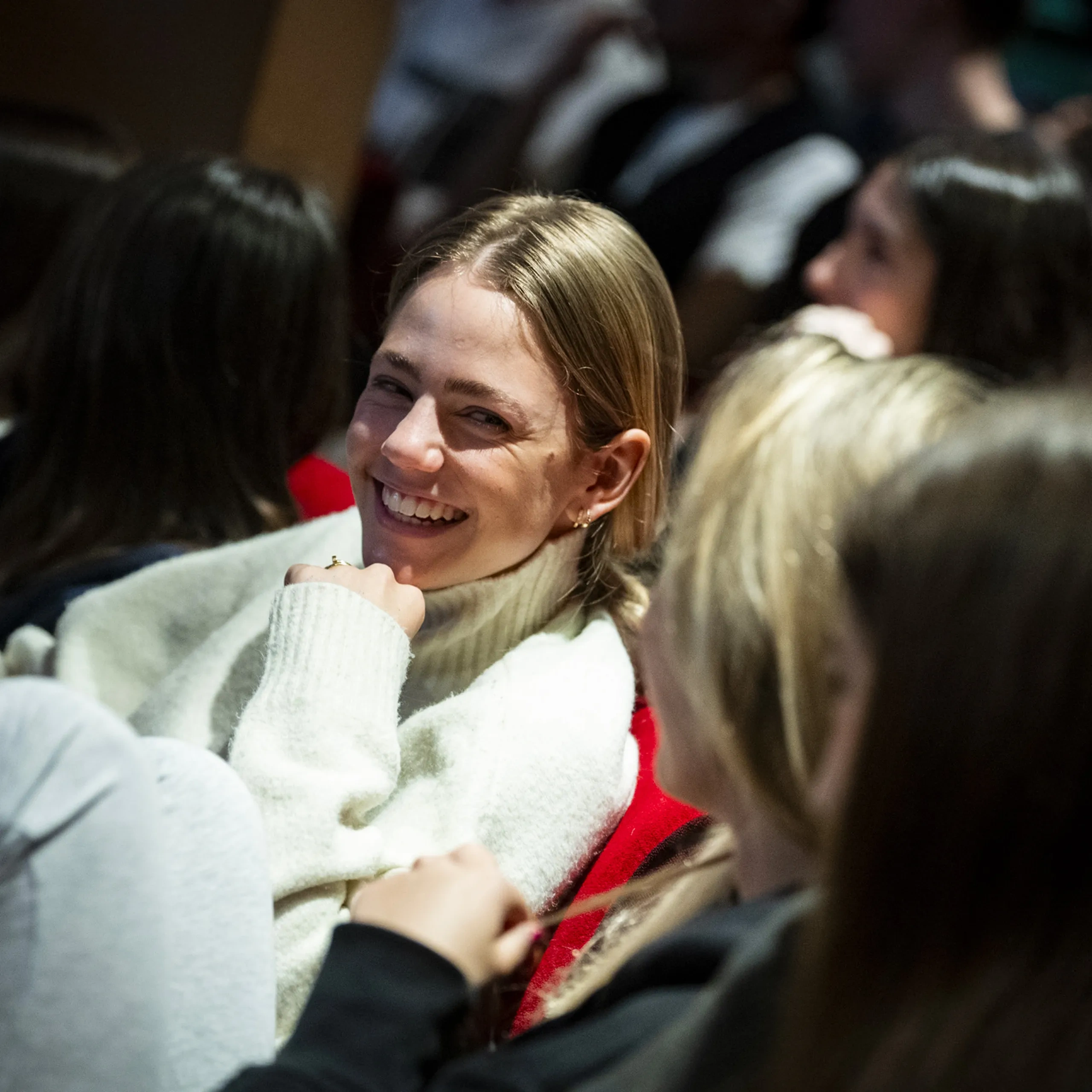 Una persona con un maglione leggero sorride mentre è seduta tra il pubblico, interagendo con le persone vicine in un ambiente poco illuminato.