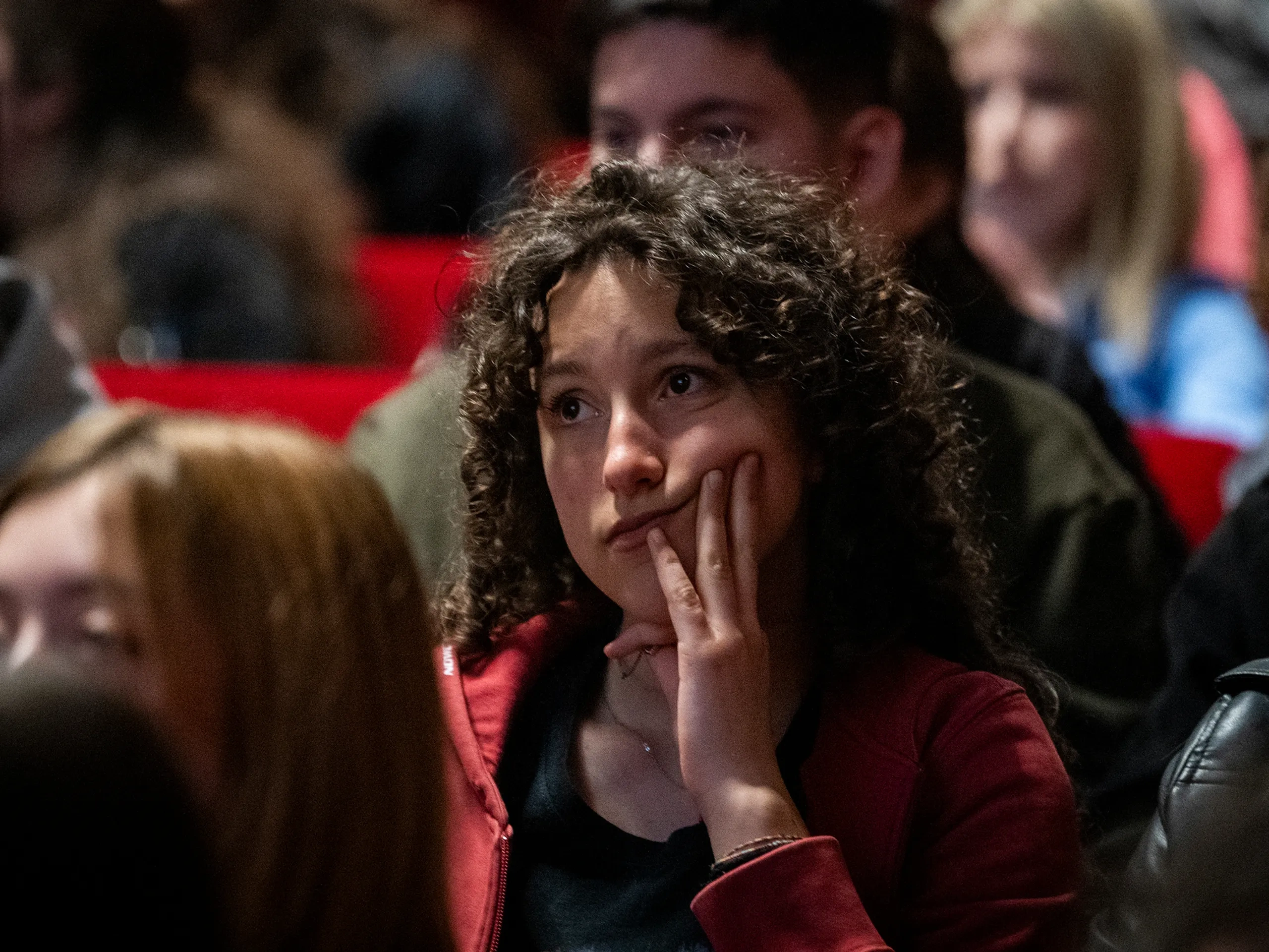 A person with curly hair in a red jacket sits in a crowd, resting their face on their hand, appearing thoughtful or contemplative.