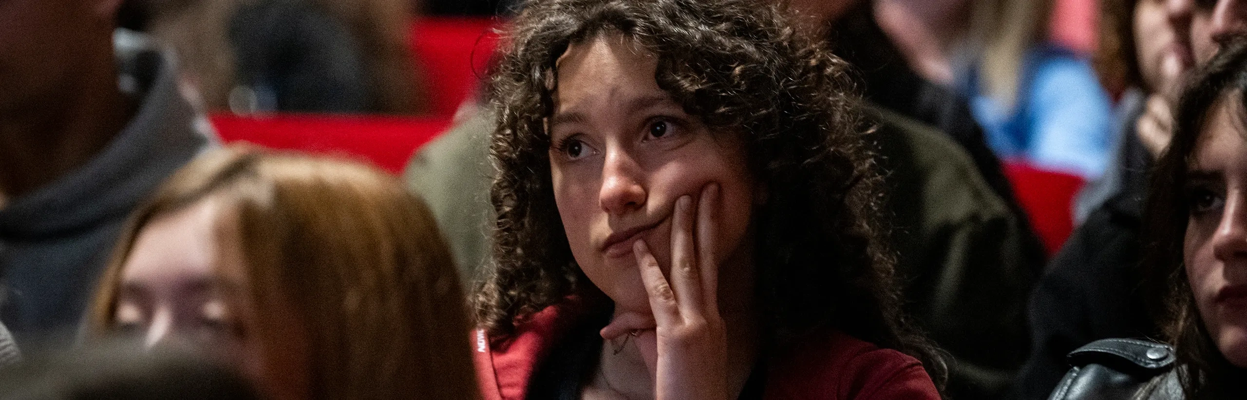 Un giovane con i capelli ricci e una giacca rossa è seduto in una stanza affollata, con il mento appoggiato sulla mano, dall'aria pensierosa.