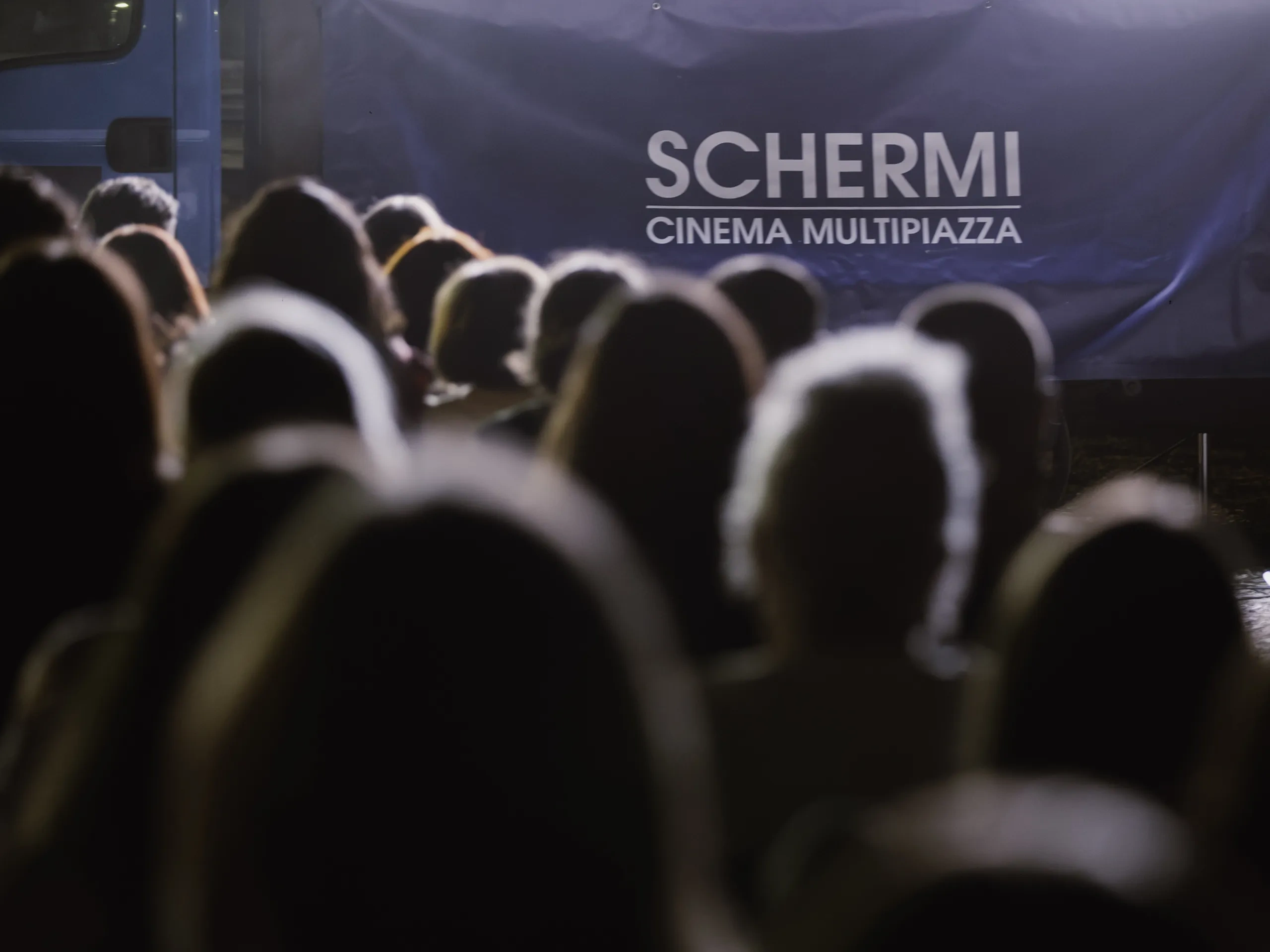 Una folla di persone rivolta verso un cartellone blu con la scritta "SCHERMI CINEMA MULTIPIAZZA", con un'illuminazione soffusa sullo sfondo.
