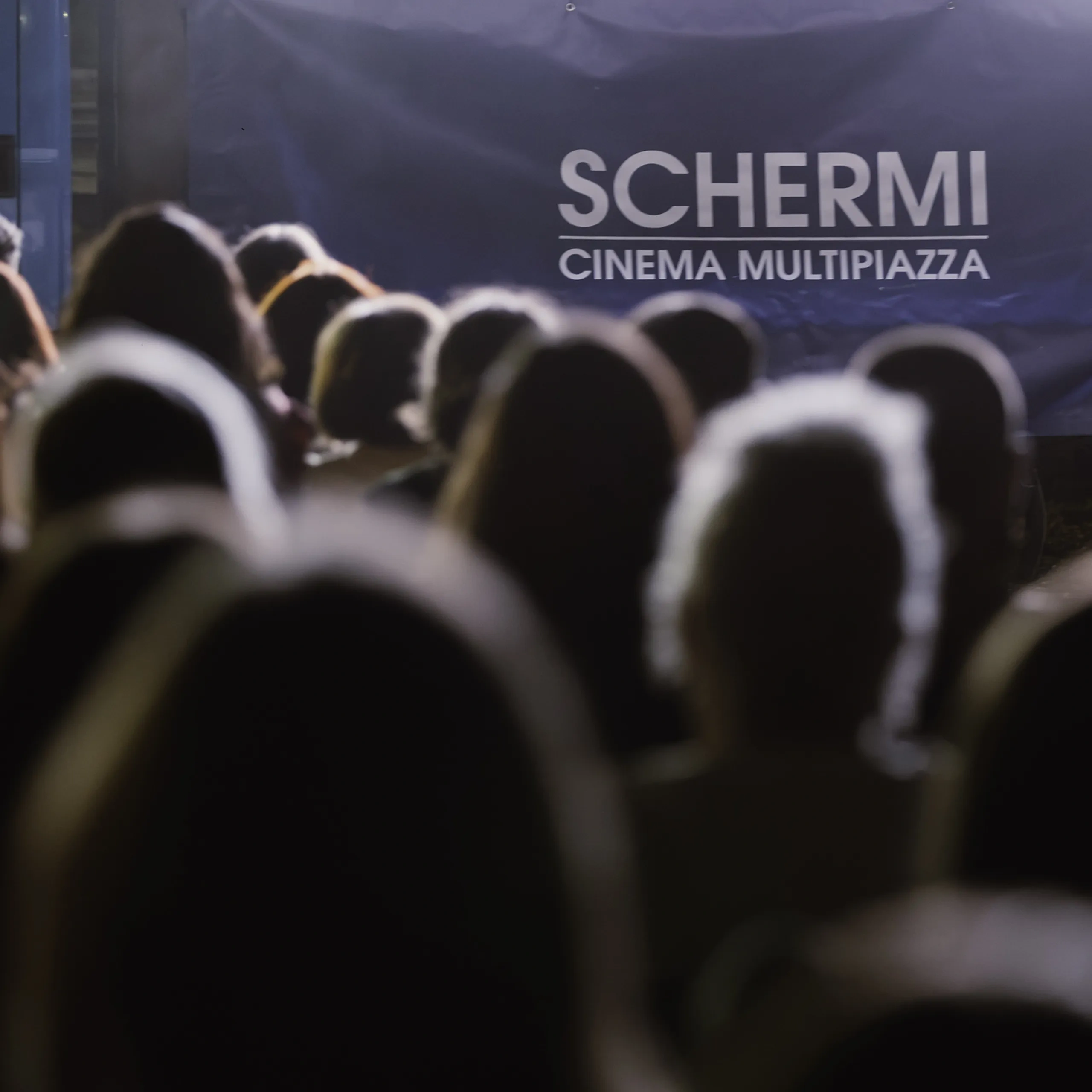 Una folla di persone rivolta verso un cartellone blu con la scritta "SCHERMI CINEMA MULTIPIAZZA", con un'illuminazione soffusa sullo sfondo.