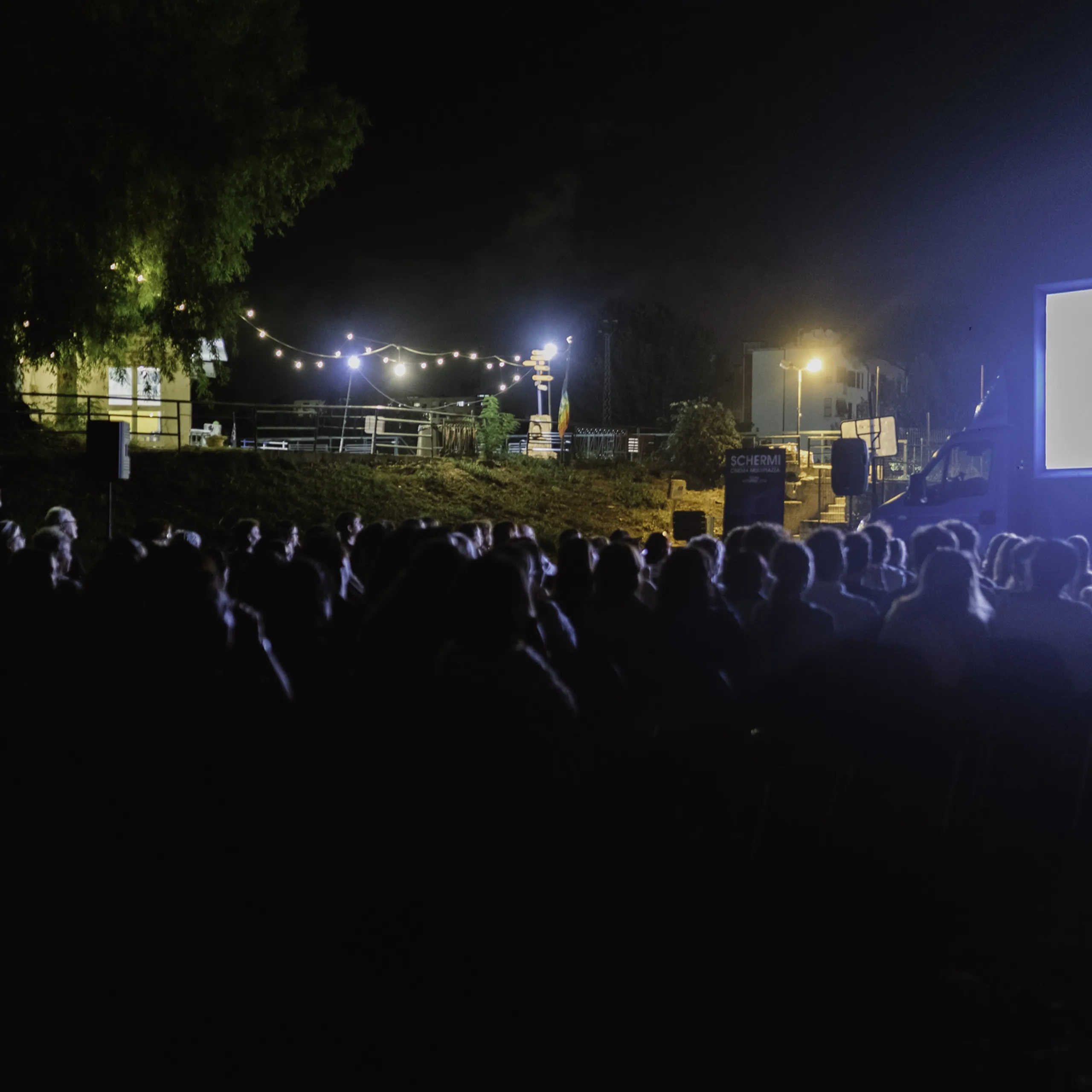 Una proiezione cinematografica all'aperto, di notte, con il pubblico seduto davanti a un grande schermo. Delle catene di luci illuminano lo sfondo.