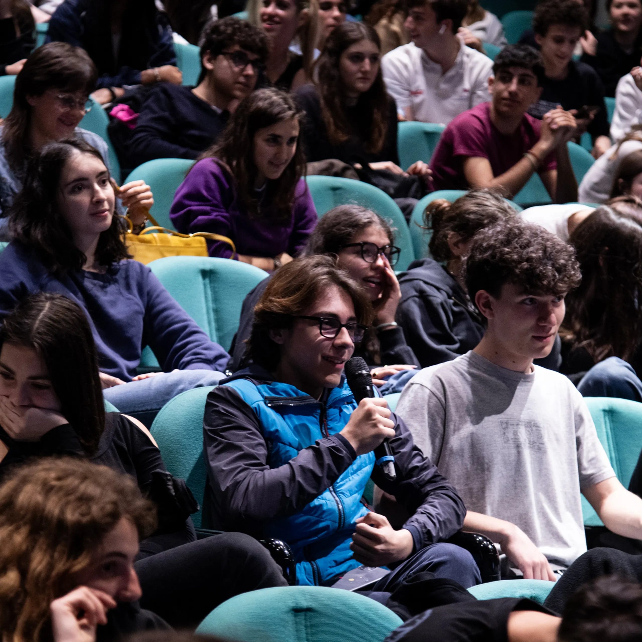 Un folto gruppo di studenti seduti in un auditorium, uno dei quali con un microfono in mano. Sembrano coinvolti e attenti.