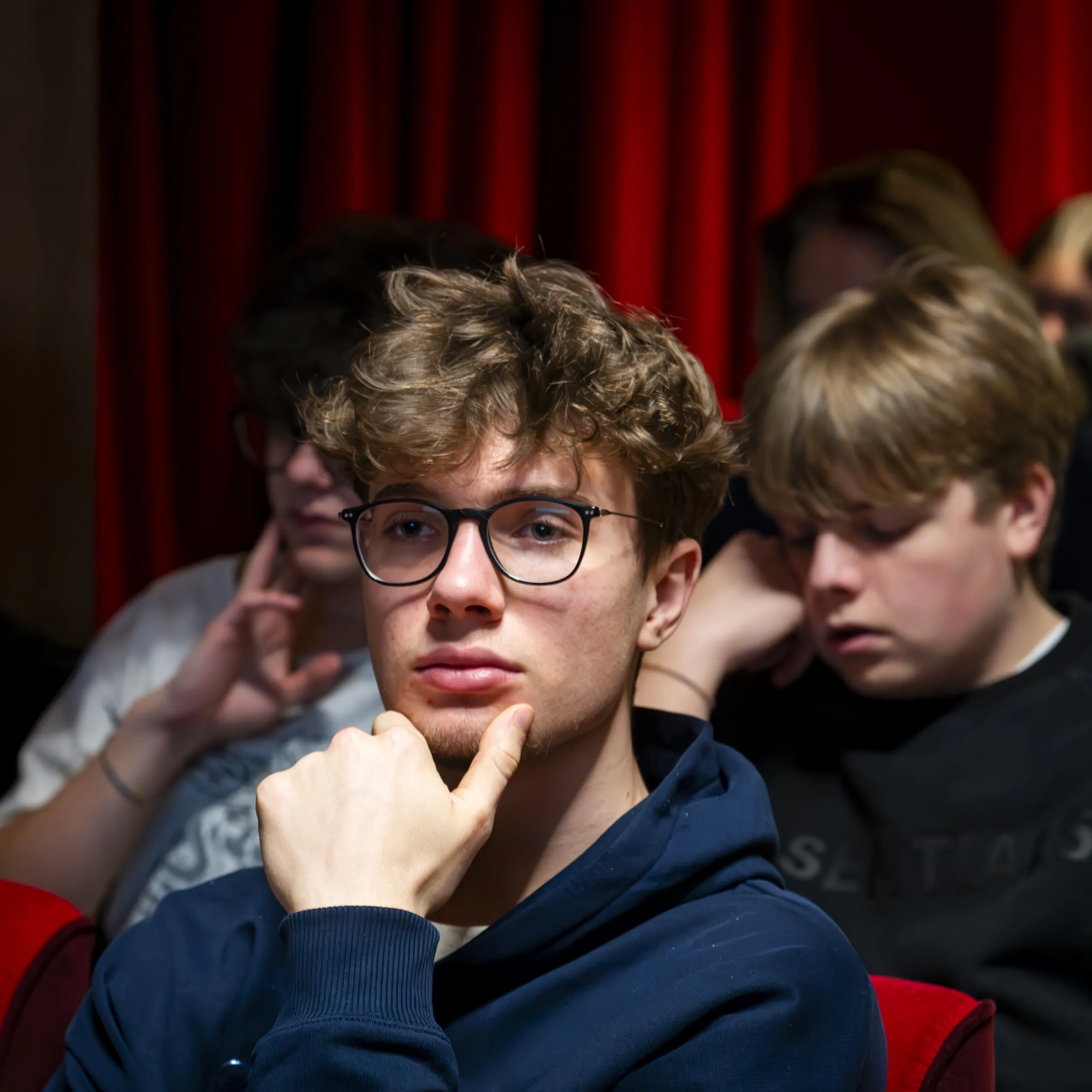 Un giovane con gli occhiali e i capelli ricci è seduto pensieroso in un teatro, circondato da altri spettatori seduti su poltrone rosse.