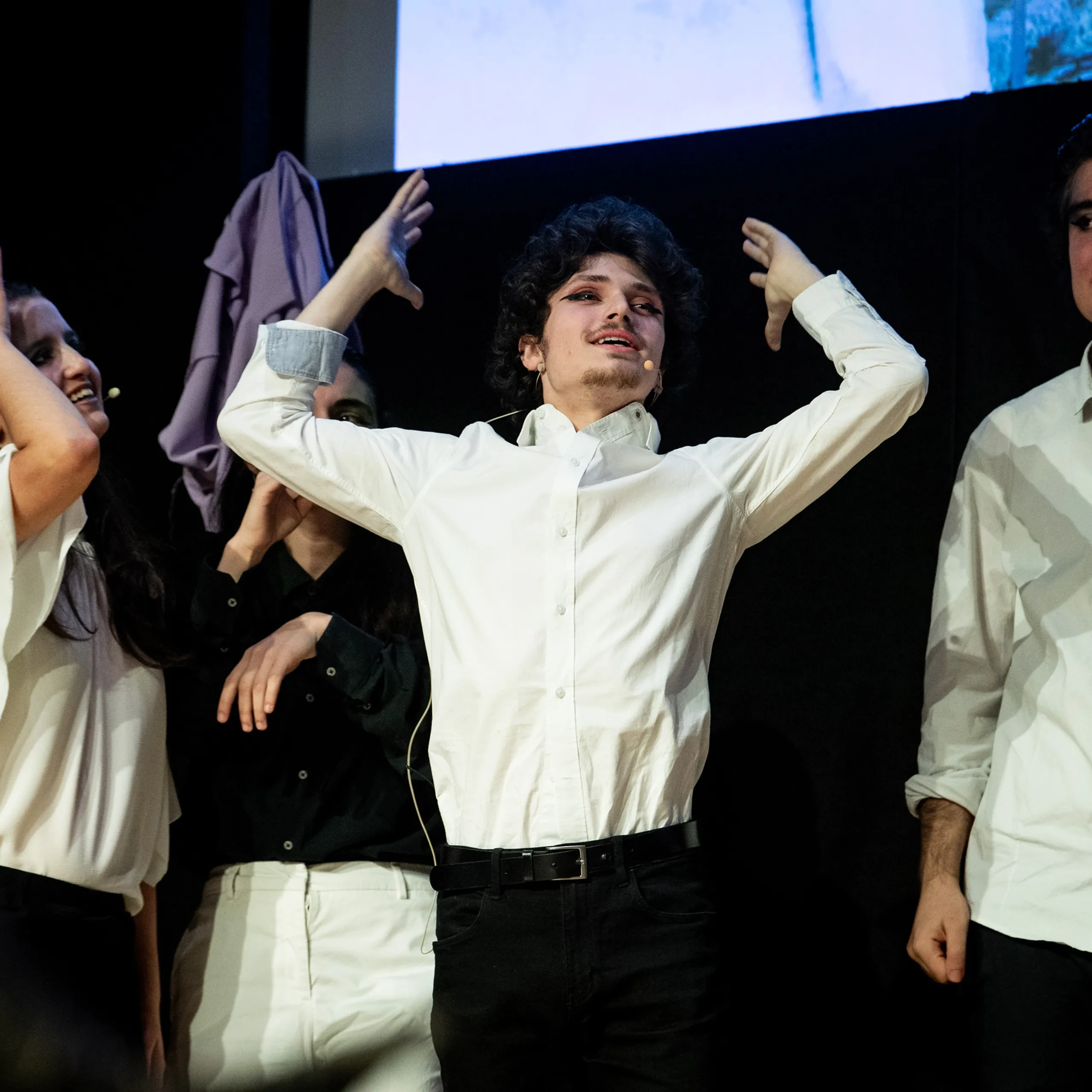 Quattro persone sul palco indossano camicie bianche. Una gesticola con le braccia alzate, mentre le altre guardano e sorridono, indicando una rappresentazione teatrale.