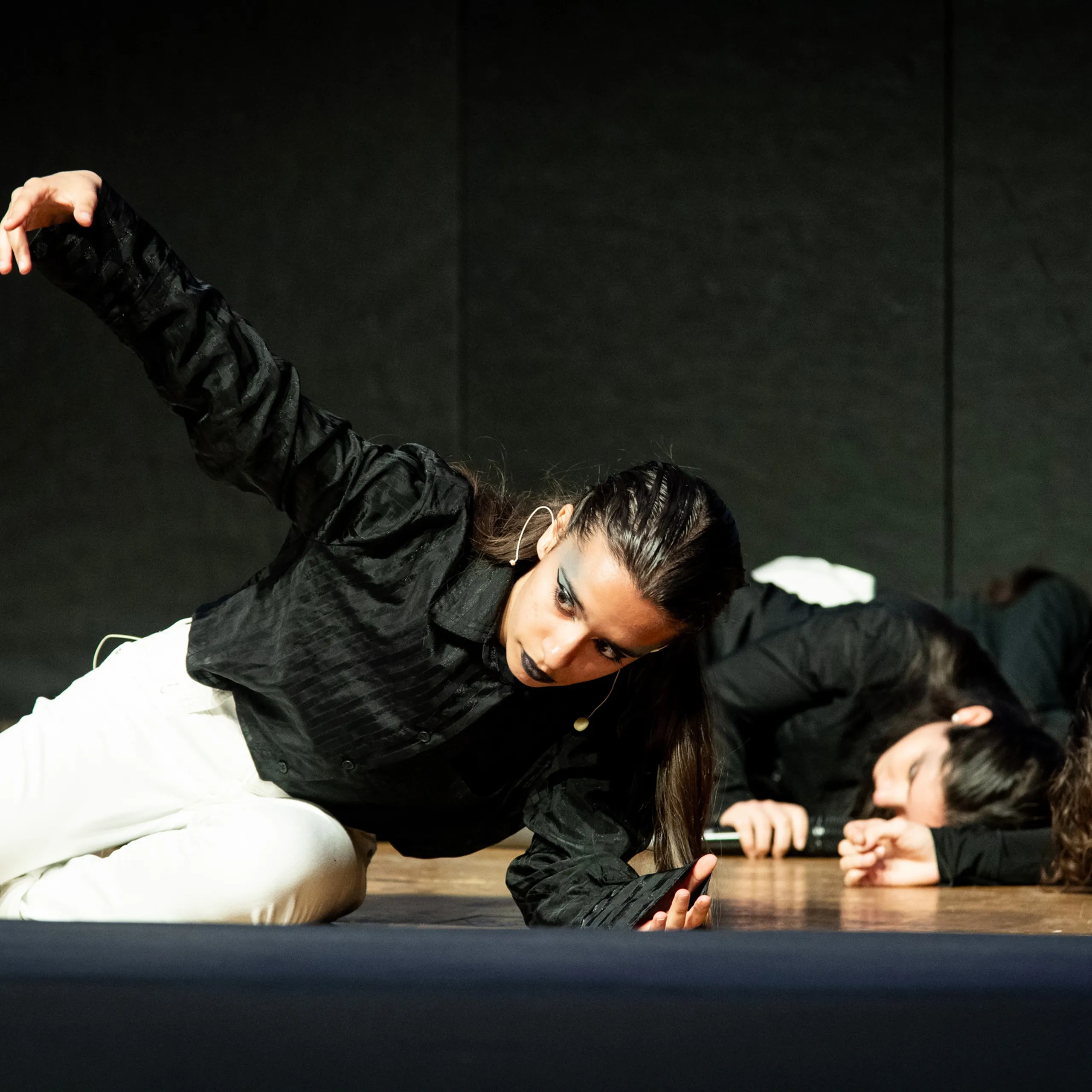 Ballerini in top neri e pantaloni bianchi si esibiscono in modo espressivo su un palco scarsamente illuminato, con uno di loro che allunga un braccio con grazia.