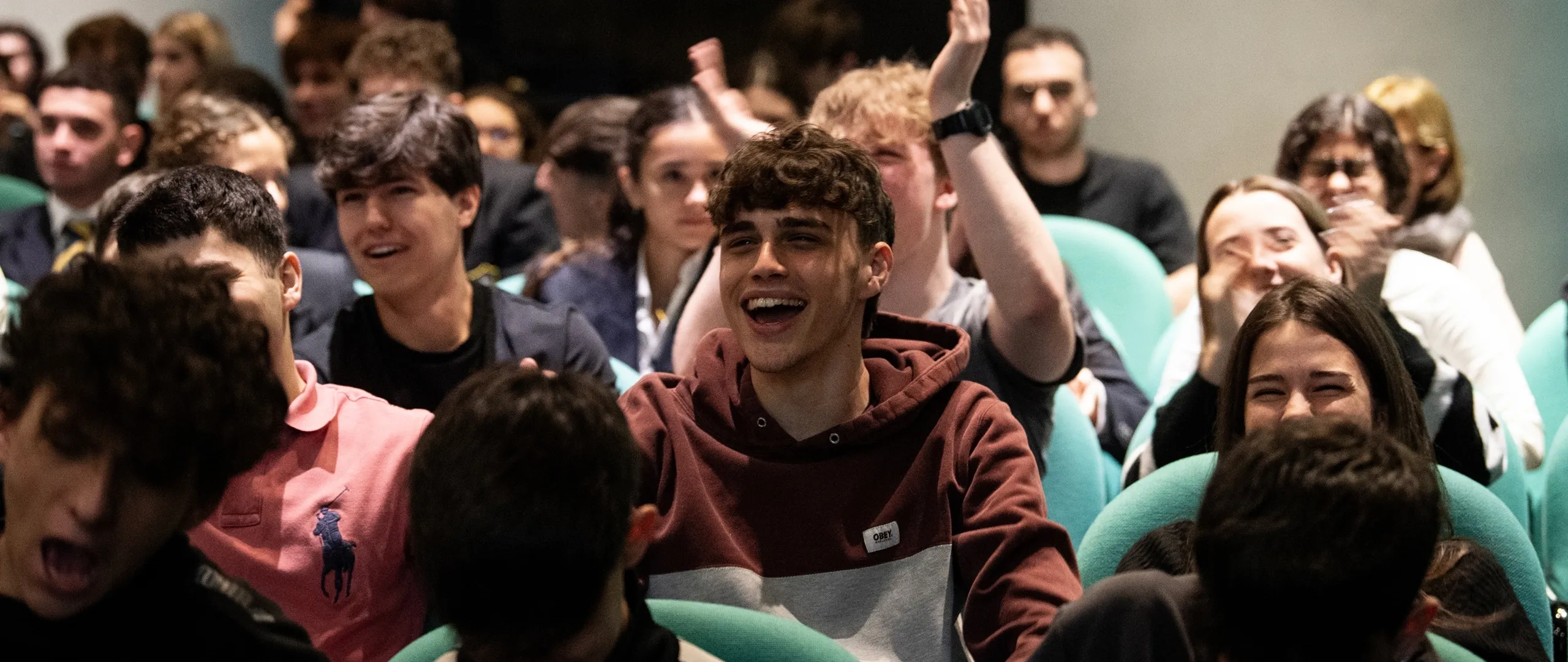 A group of people seated in a theater, smiling and clapping, with one person at the center in a maroon hoodie.