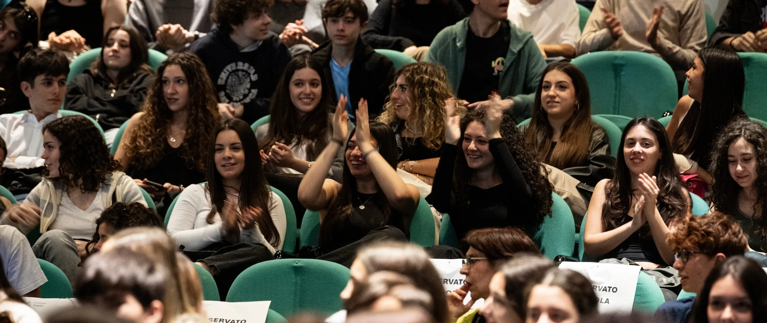 Un folto gruppo di persone sedute in una sala simile a un teatro, alcune delle quali applaudono mentre altre conversano. Sedili rivestiti in tessuto verde.