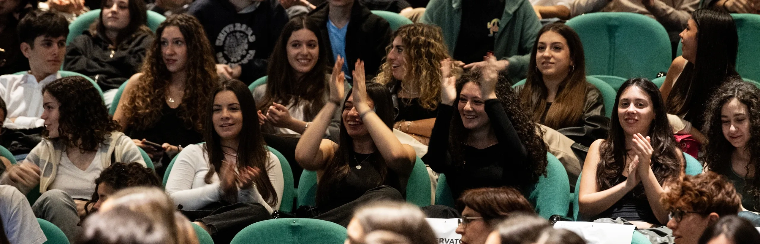 A large group of people seated in a theater, many clapping and smiling, suggesting they are watching a performance or presentation.