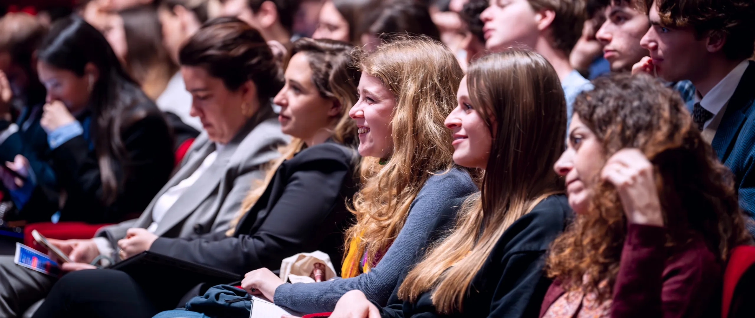 Audience seated in a theater, focused on an event. People of varying ages are engaged, seated in red chairs, with a blurred background.
