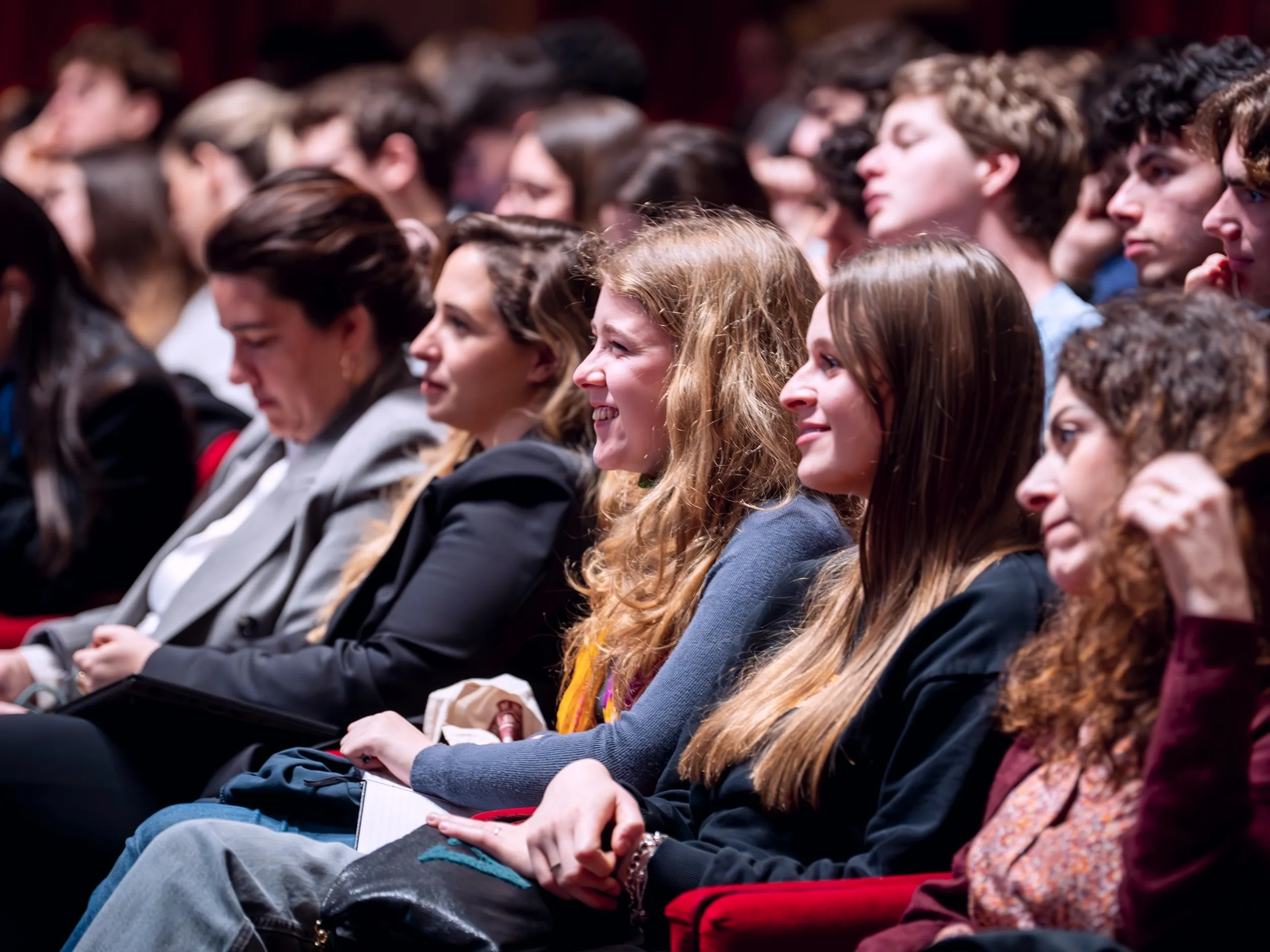 Spettatori seduti in un teatro, che guardano attentamente uno spettacolo o una presentazione, con espressioni concentrate e coinvolte.