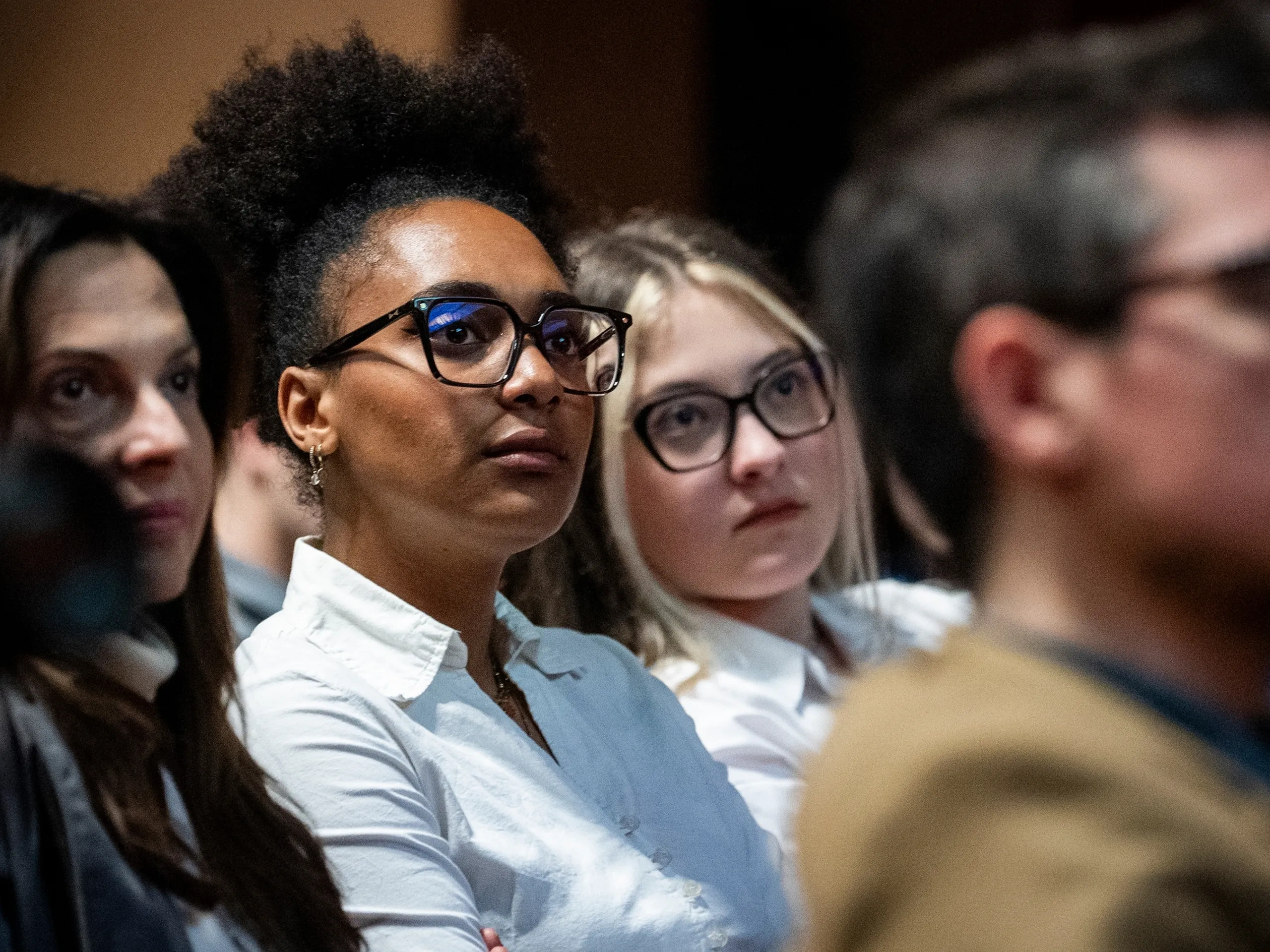 A diverse group of people seated attentively, with a woman wearing glasses and a white shirt in focus.