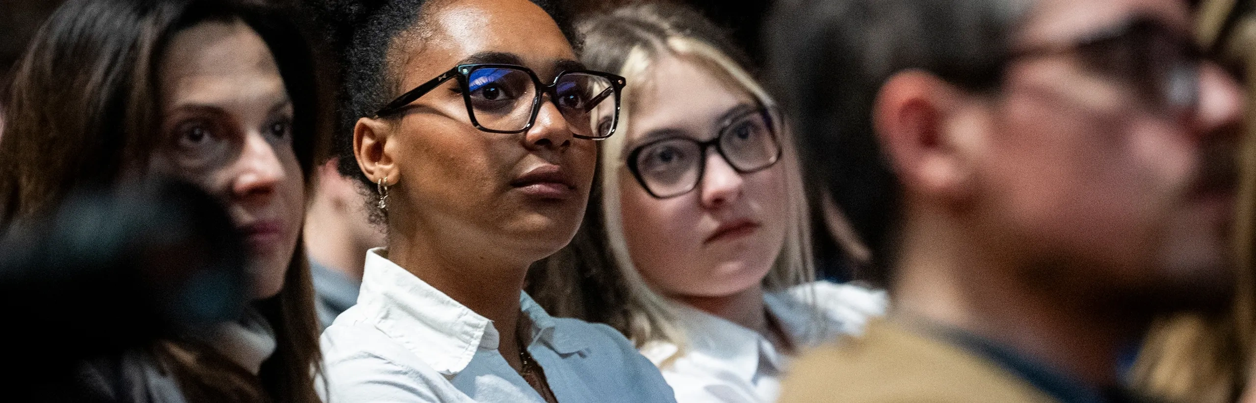 A diverse group of people attentively listening in an indoor setting, with a woman in glasses and a white shirt in focus.