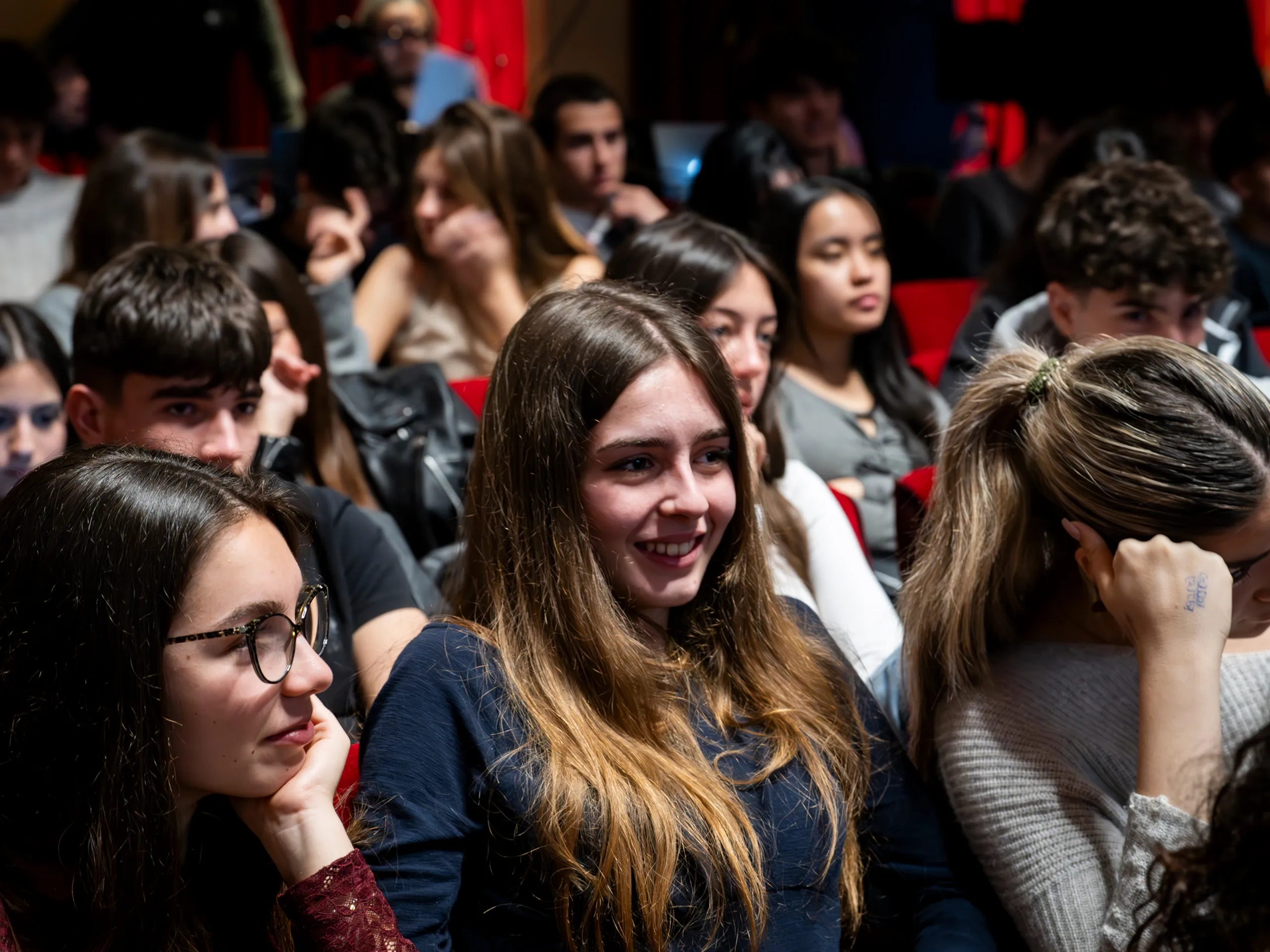 Pubblico di giovani seduti, che guardano attentamente un evento, con particolare attenzione a una donna sorridente al centro.