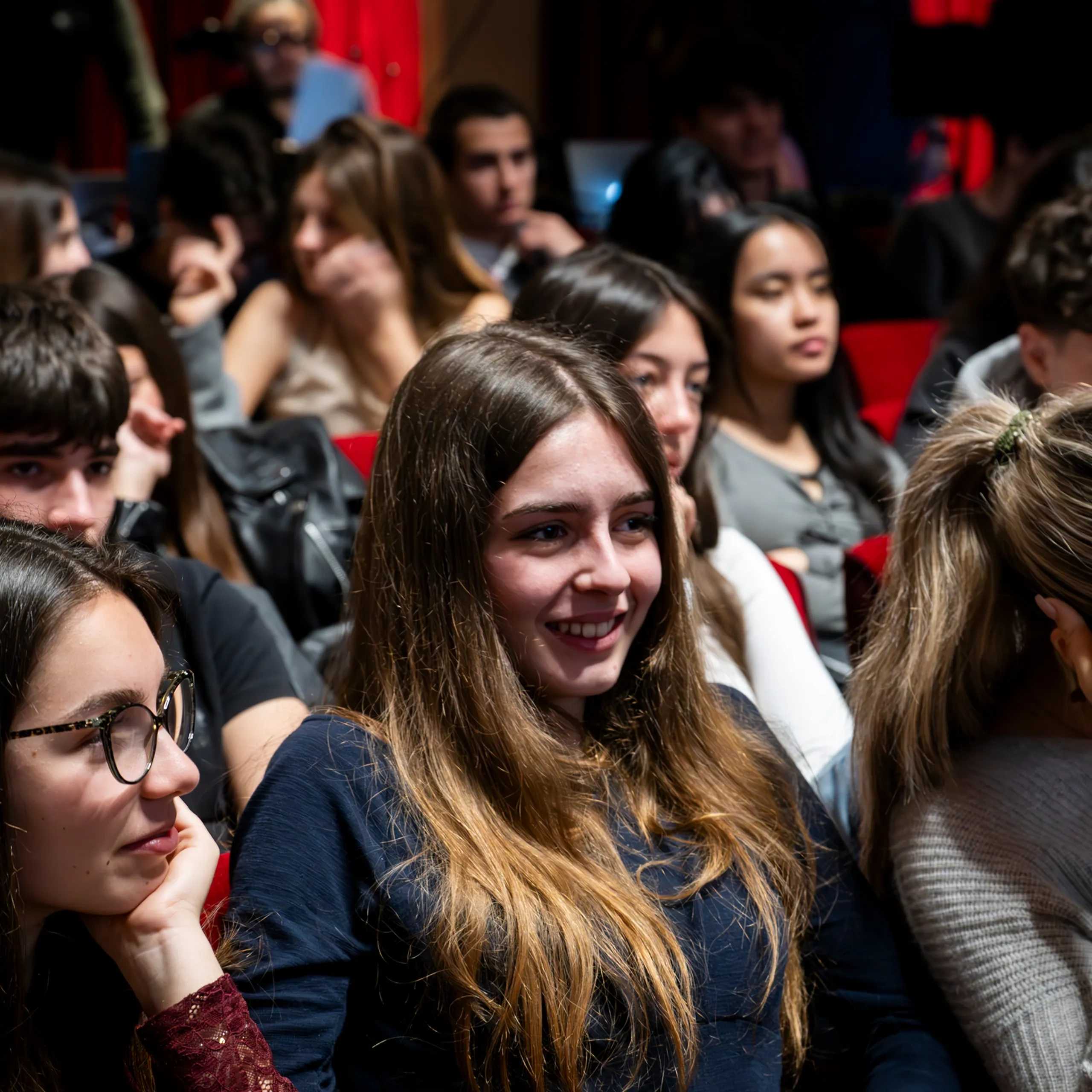 Pubblico di giovani seduti, che guardano attentamente un evento, con particolare attenzione a una donna sorridente al centro.