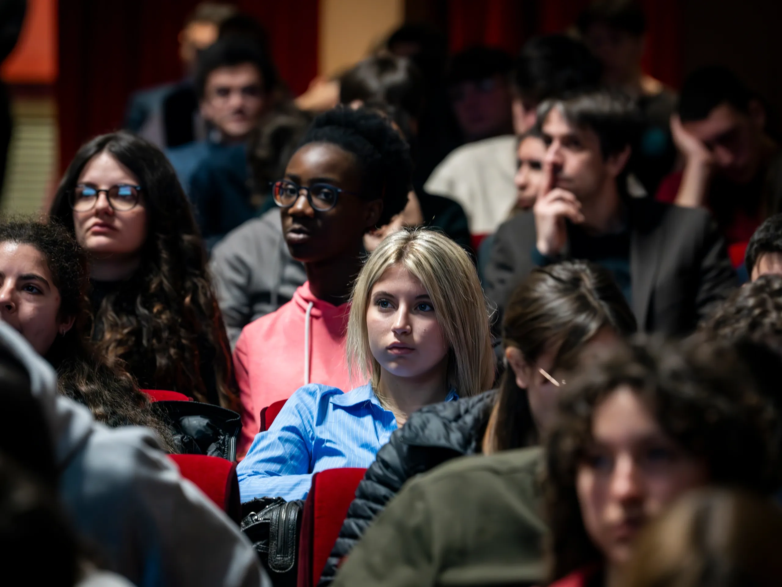 Un gruppo eterogeneo di giovani seduti in un auditorium che ascoltano attentamente una presentazione o un evento.