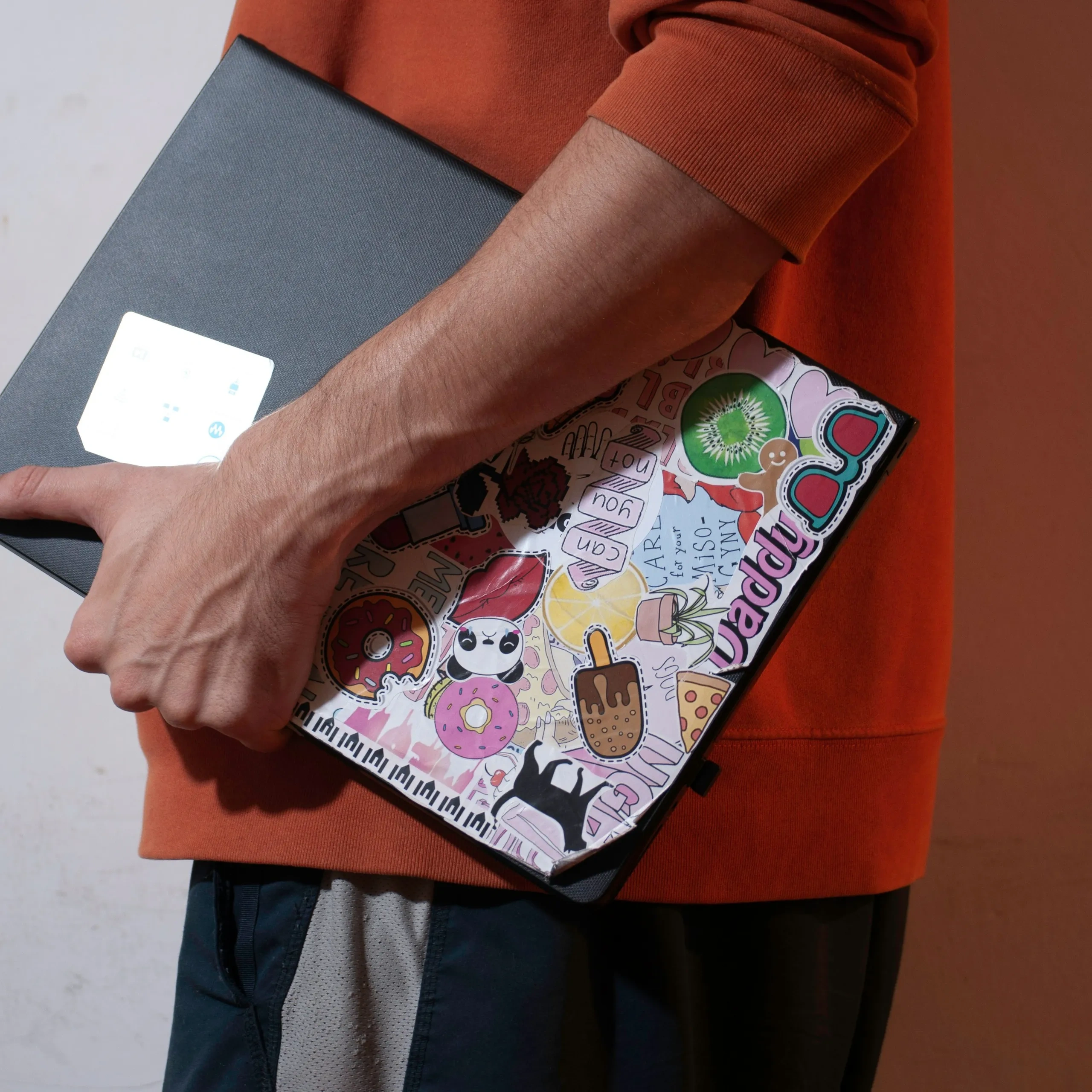 Persona con una felpa arancione che tiene in mano un laptop con adesivi colorati e originali su uno sfondo semplice.