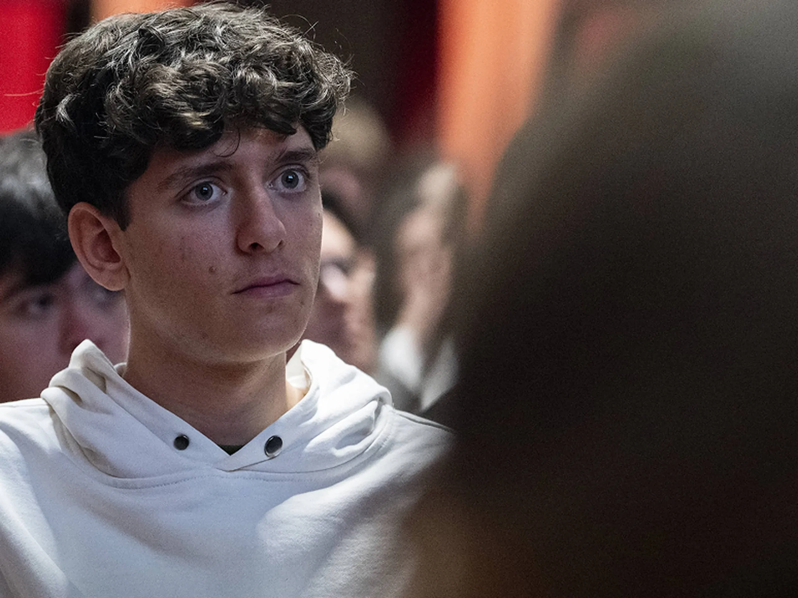 Un giovane con i capelli ricci e una felpa bianca con cappuccio ascolta attentamente in un ambiente interno affollato.