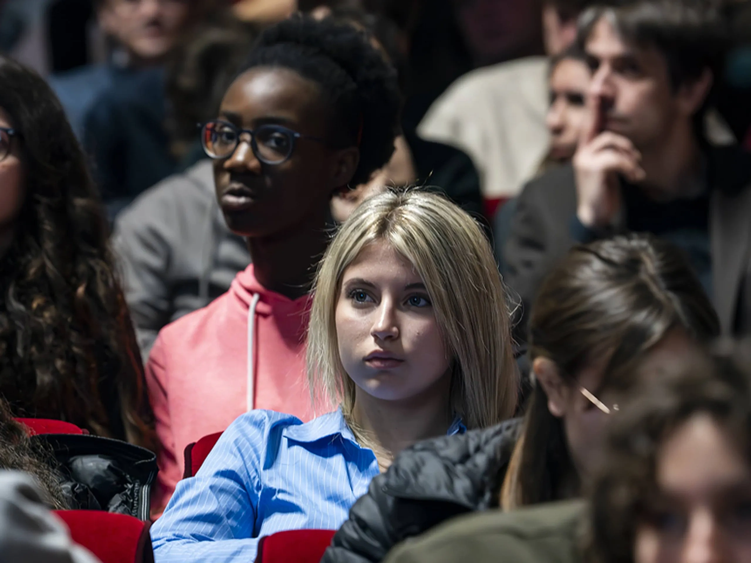Pubblico composto da persone diverse sedute, che guardano attentamente un evento in un auditorium poco illuminato.