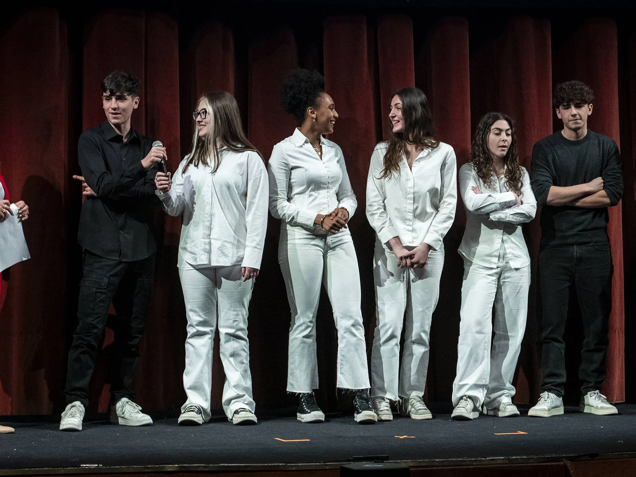A group of seven casually dressed individuals stand on a stage, some holding papers, in front of a red curtain.