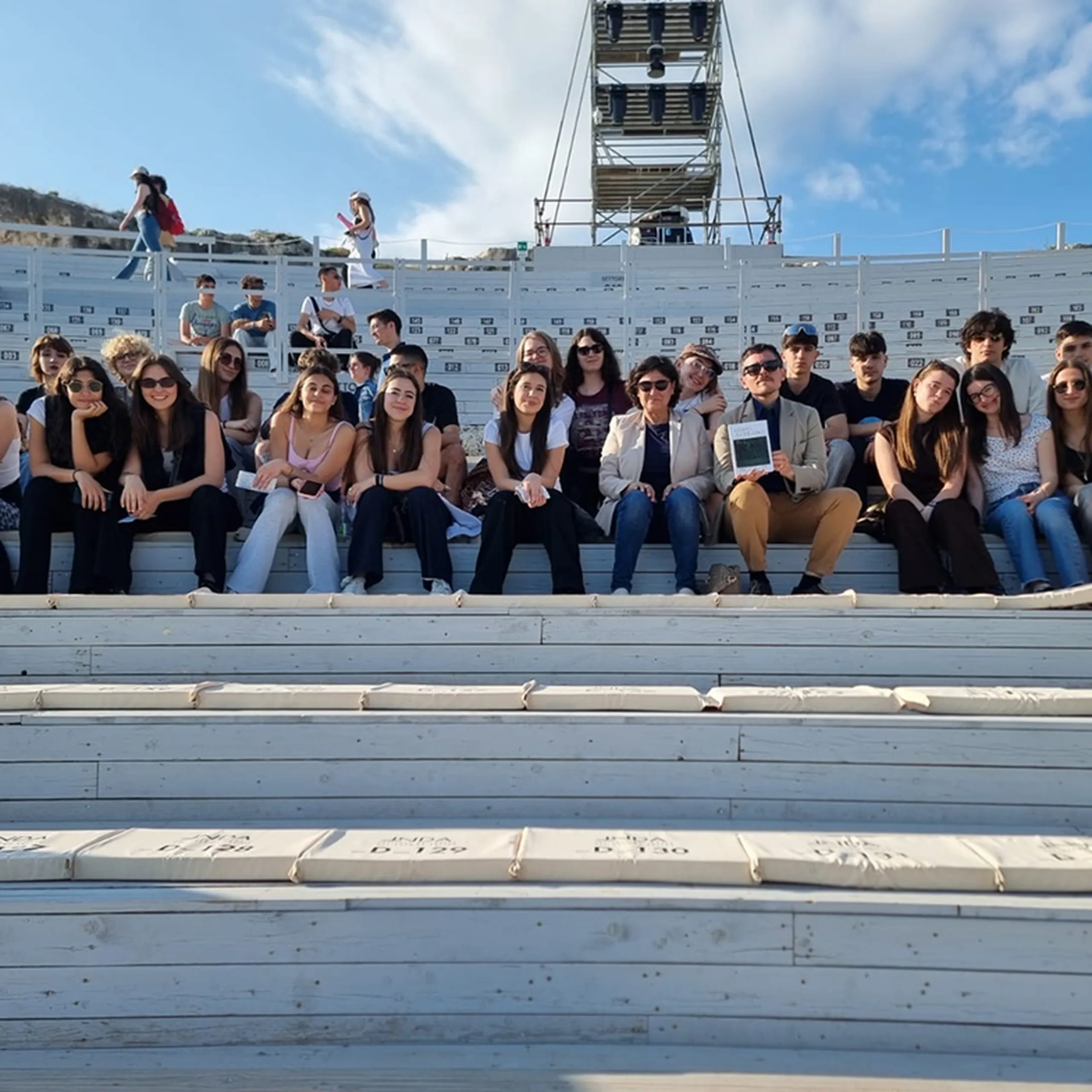 Un gruppo di persone sedute in file su gradini all'aperto. Un palco con attrezzature dietro di loro, sotto un cielo parzialmente nuvoloso.