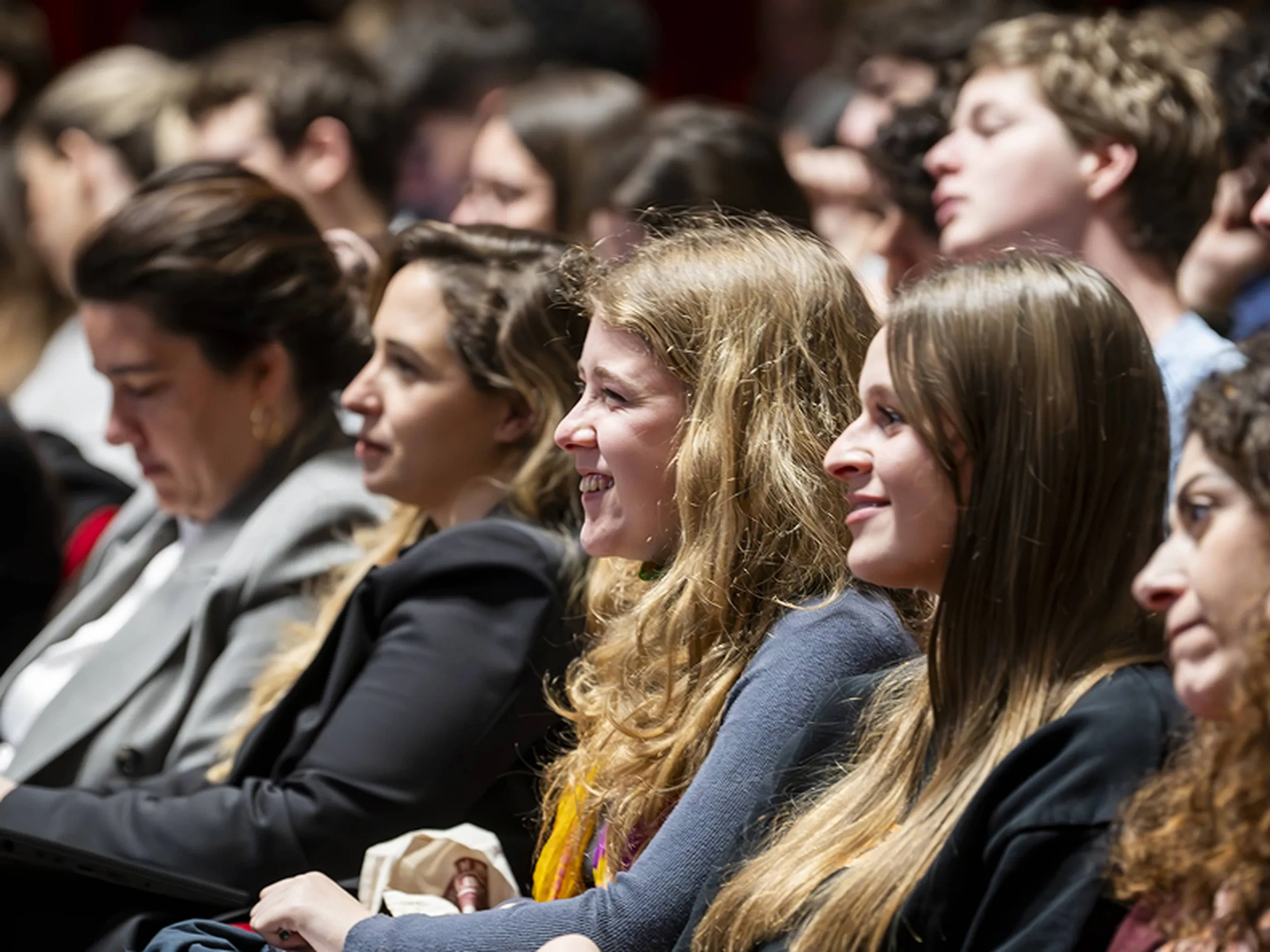 Il pubblico è seduto in un teatro, concentrato e attento, con alcune persone che sorridono. L'illuminazione è calda e mette in risalto le loro espressioni.