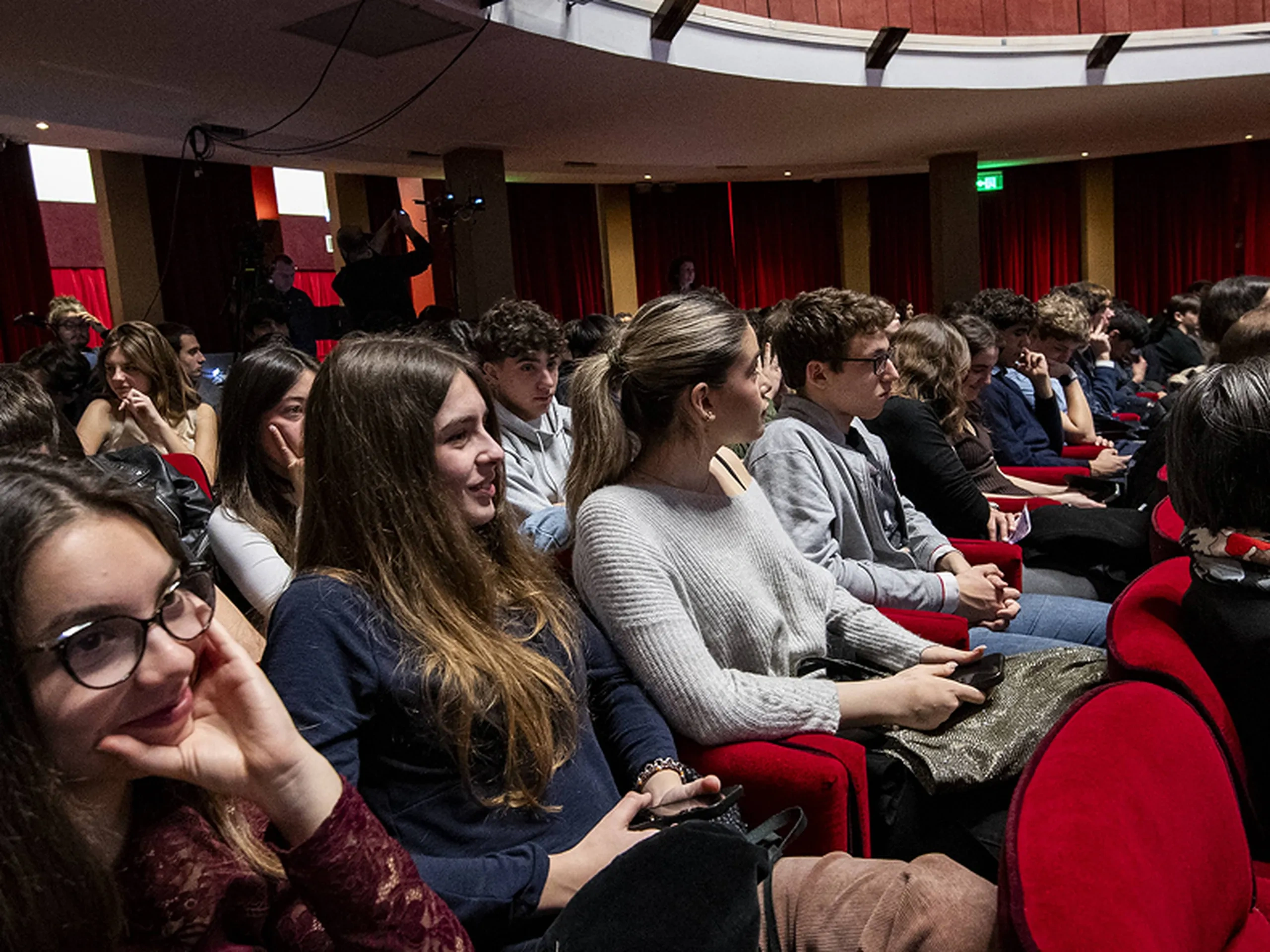 Pubblico seduto in un teatro, attento a una presentazione, con sedili rossi e luci soffuse.