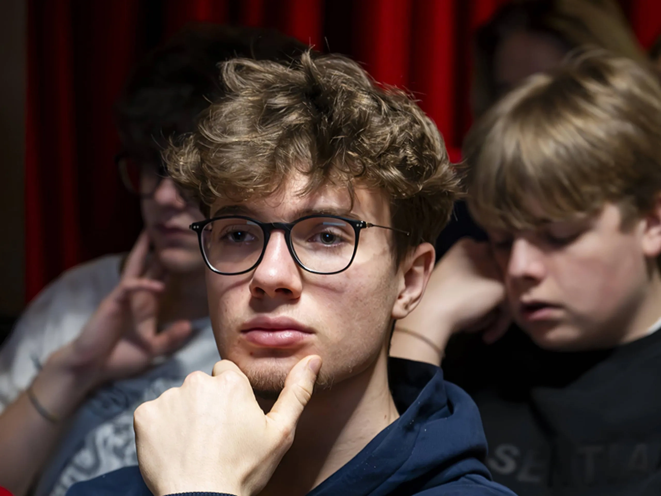 Giovane uomo con capelli ricci e occhiali, seduto in una stanza poco illuminata, che indossa una felpa con cappuccio scura, con persone sfocate sullo sfondo.