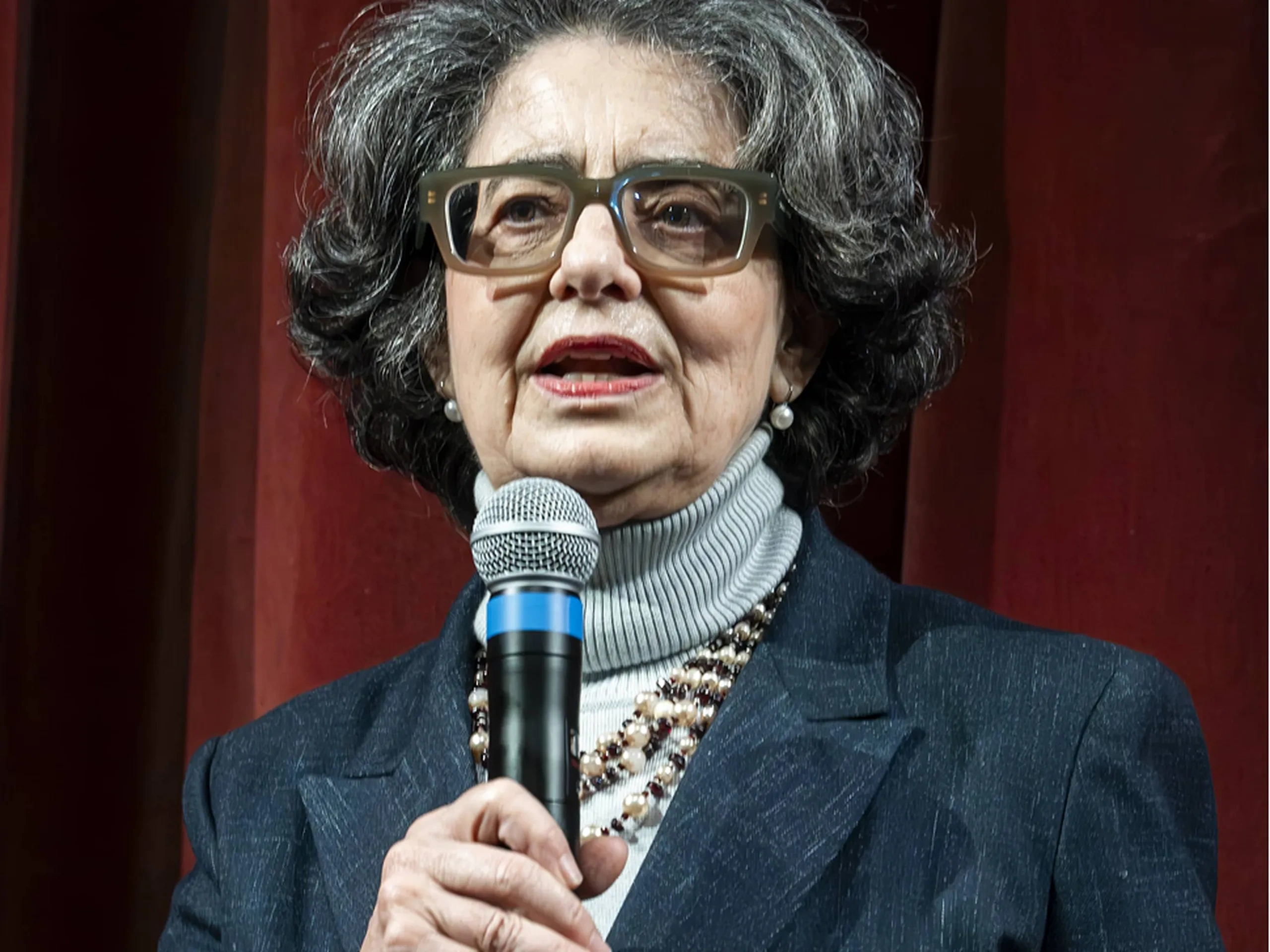 Una donna anziana con gli occhiali parla al microfono sul palco, indossando un dolcevita grigio, un blazer blu e una collana di perline, sullo sfondo di un sipario rosso.