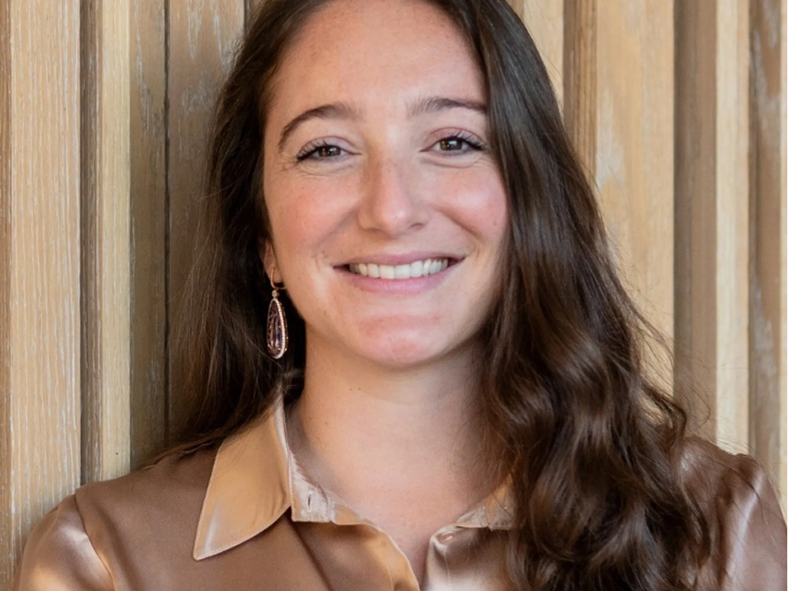 Donna sorridente, con indosso una camicetta di raso e orecchini a cerchio, in piedi su uno sfondo con texture in legno. Capelli castani sciolti e mossi.